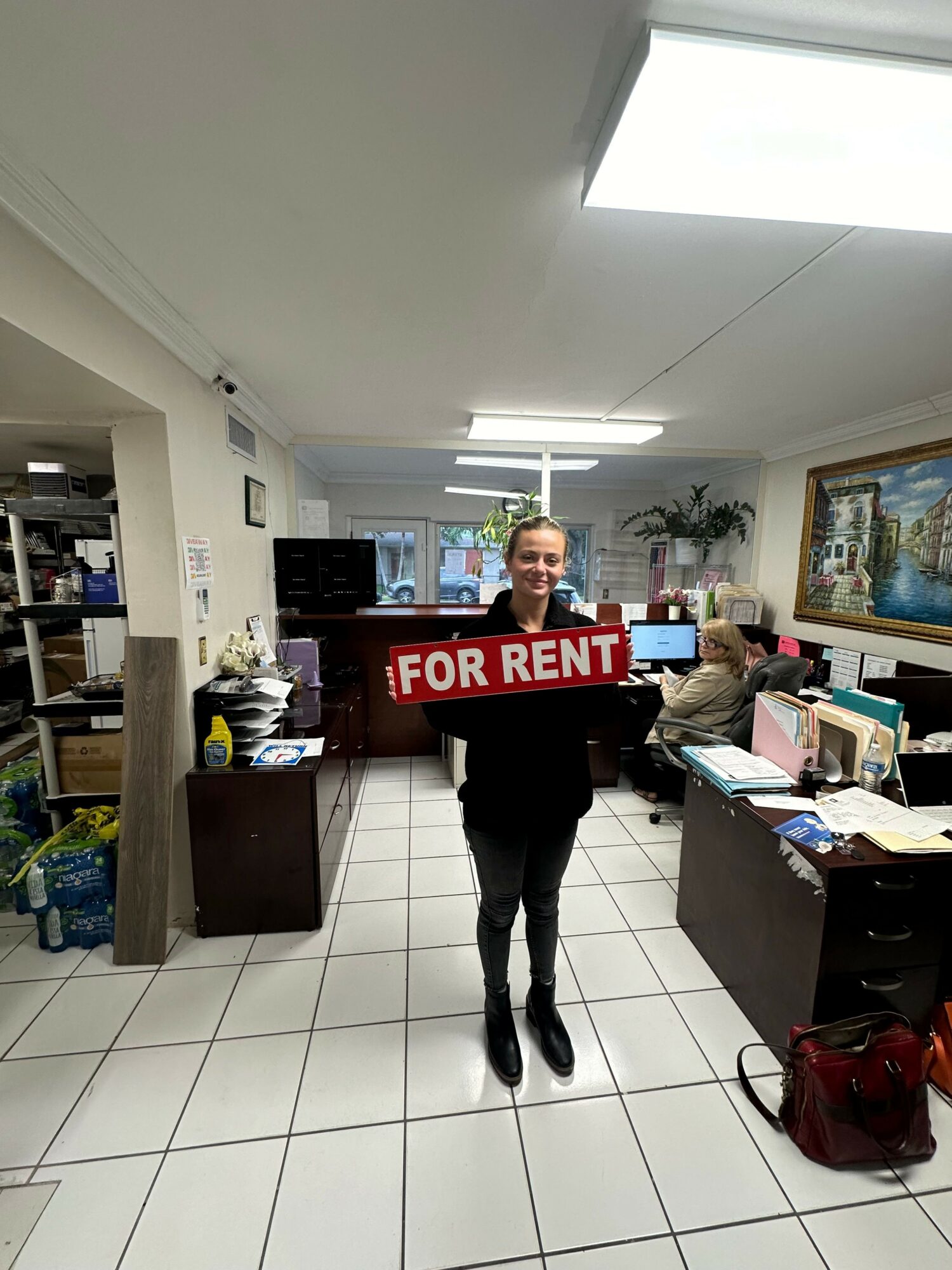 Person holding a red 'FOR RENT' sign in an office with desks and computers.