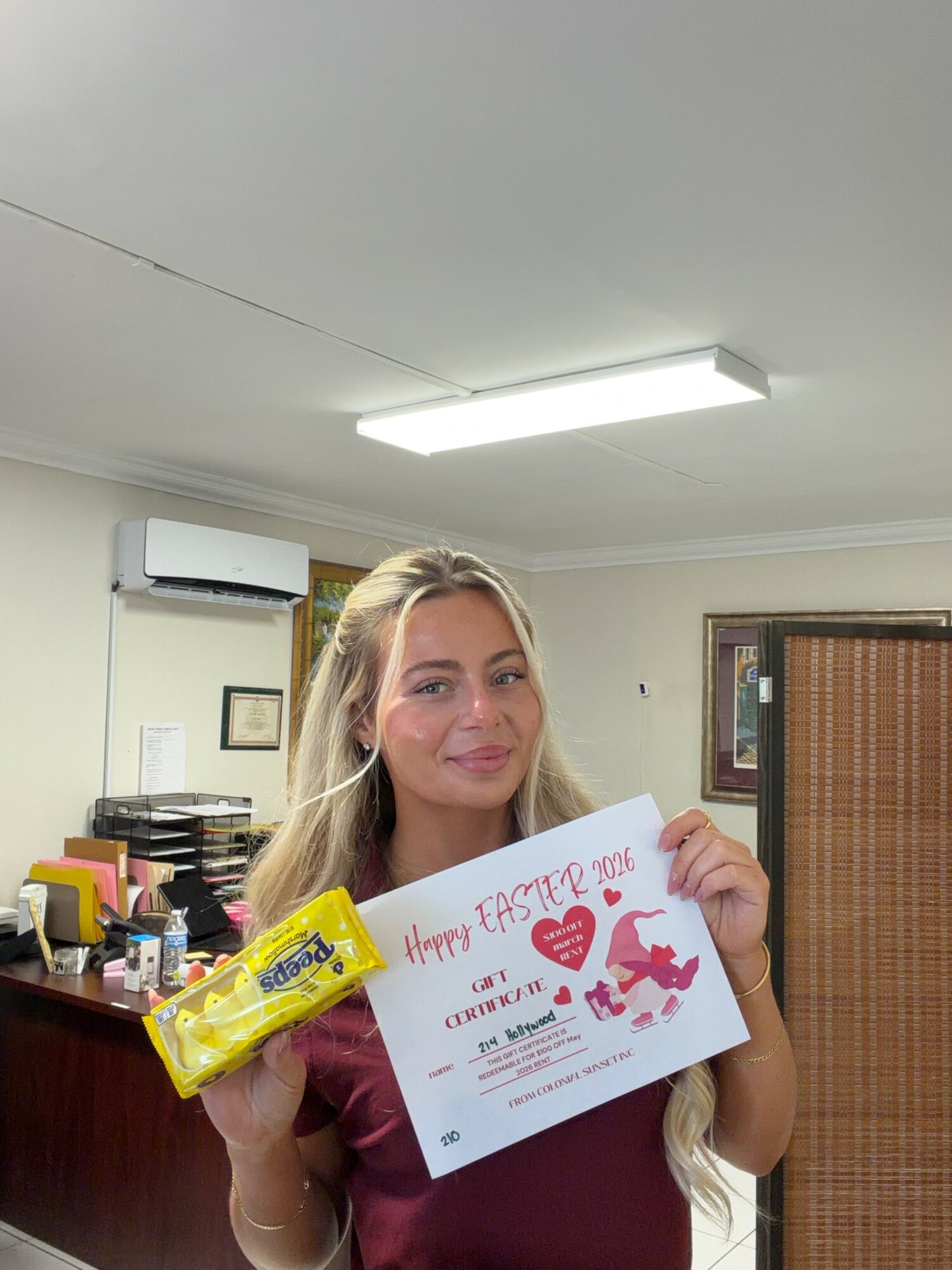 Young woman with long blonde hair holding a yellow snack and a handwritten Easter card, smiling indoors.