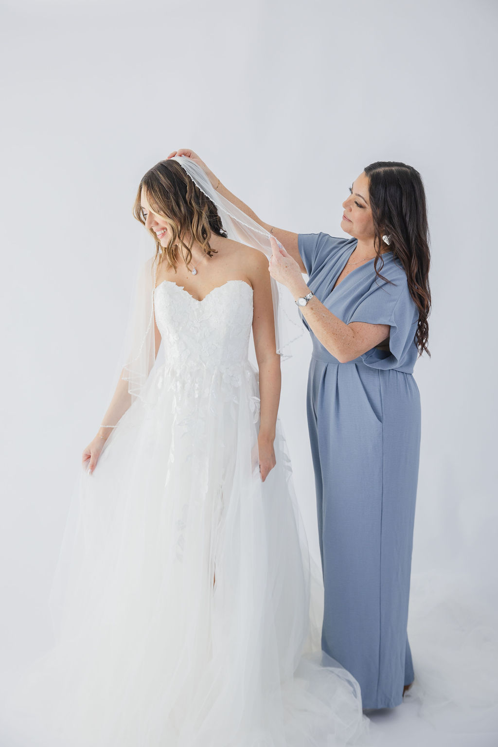 Woman in blue dress adjusting a bride's veil against a plain white background.