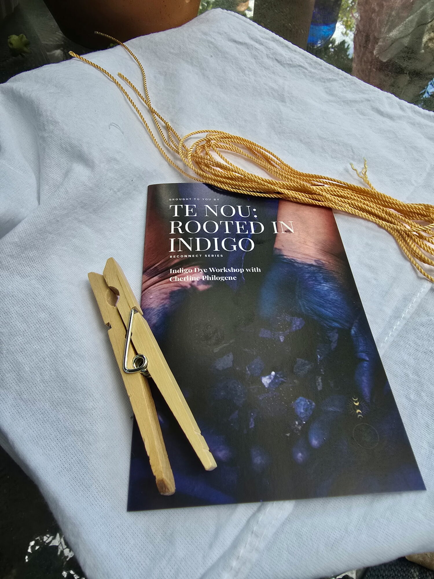 Book titled 'The Now: Rooted in Indigo' with a yellow clothespin and a bundle of gold thread on a white cloth surface.
