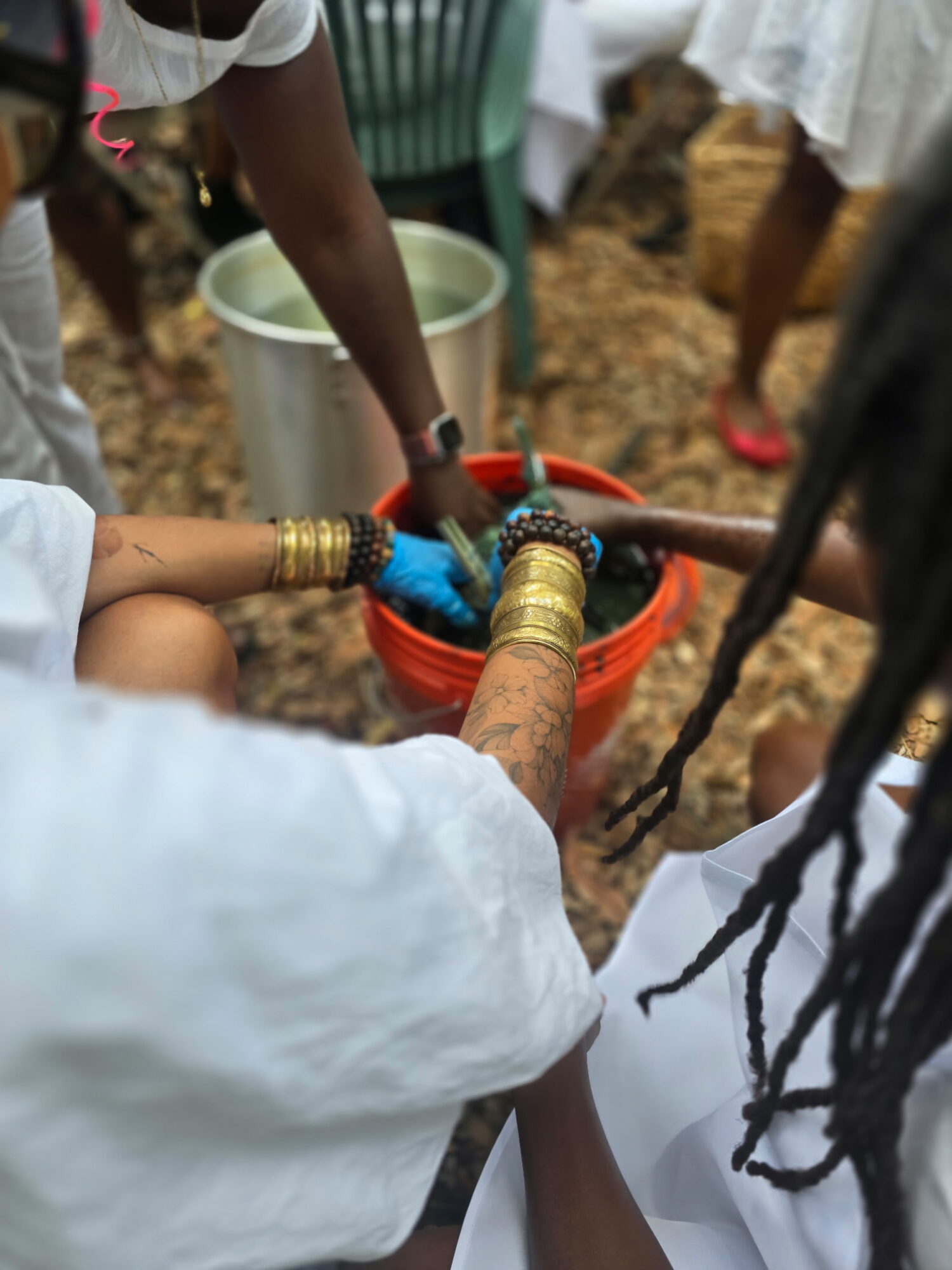 People gather around a water source, filling containers with water outdoors, with a bucket and ground visible.