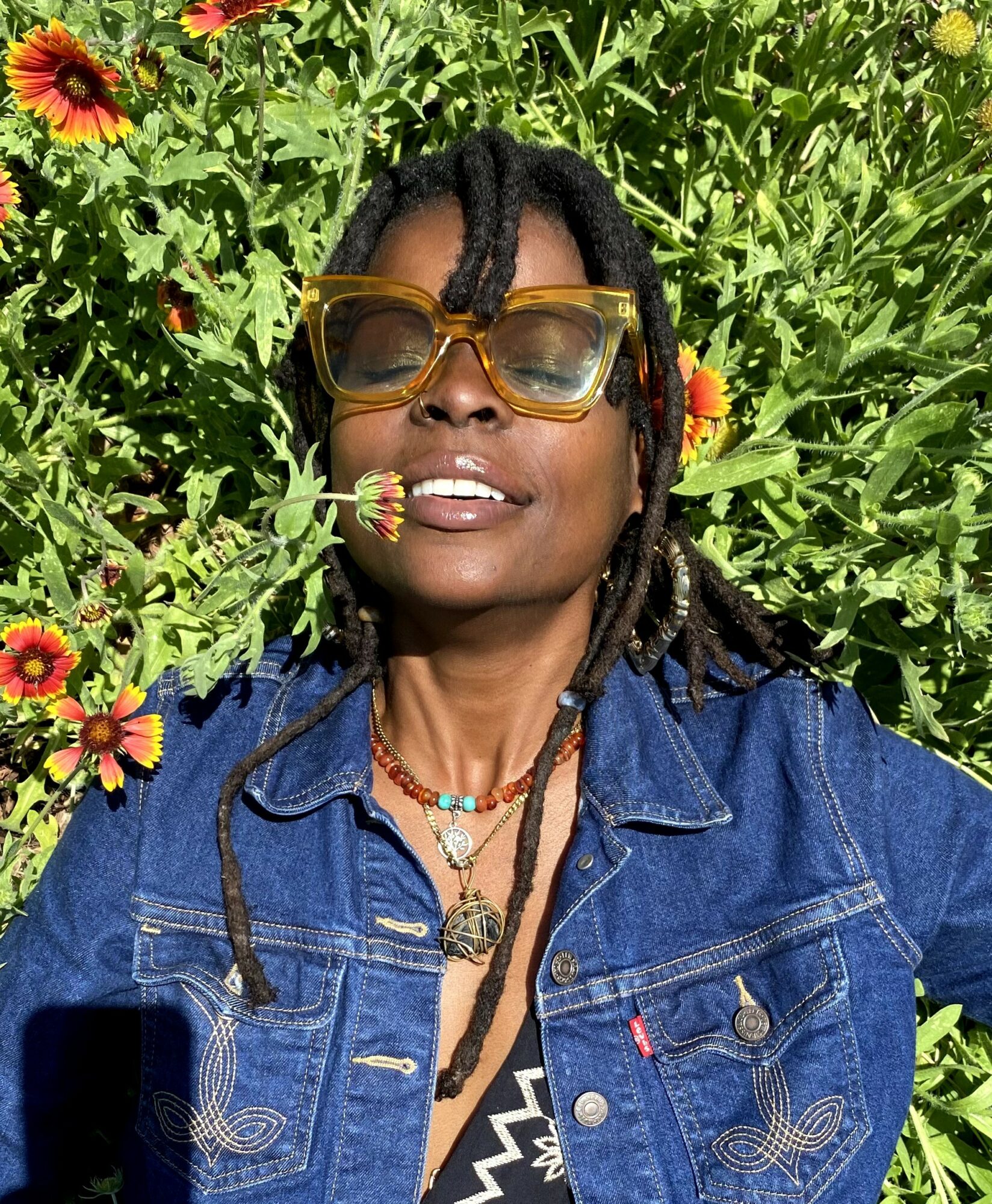 Woman with dreadlocks wearing sunglasses and denim jacket lying in a garden with colorful flowers.