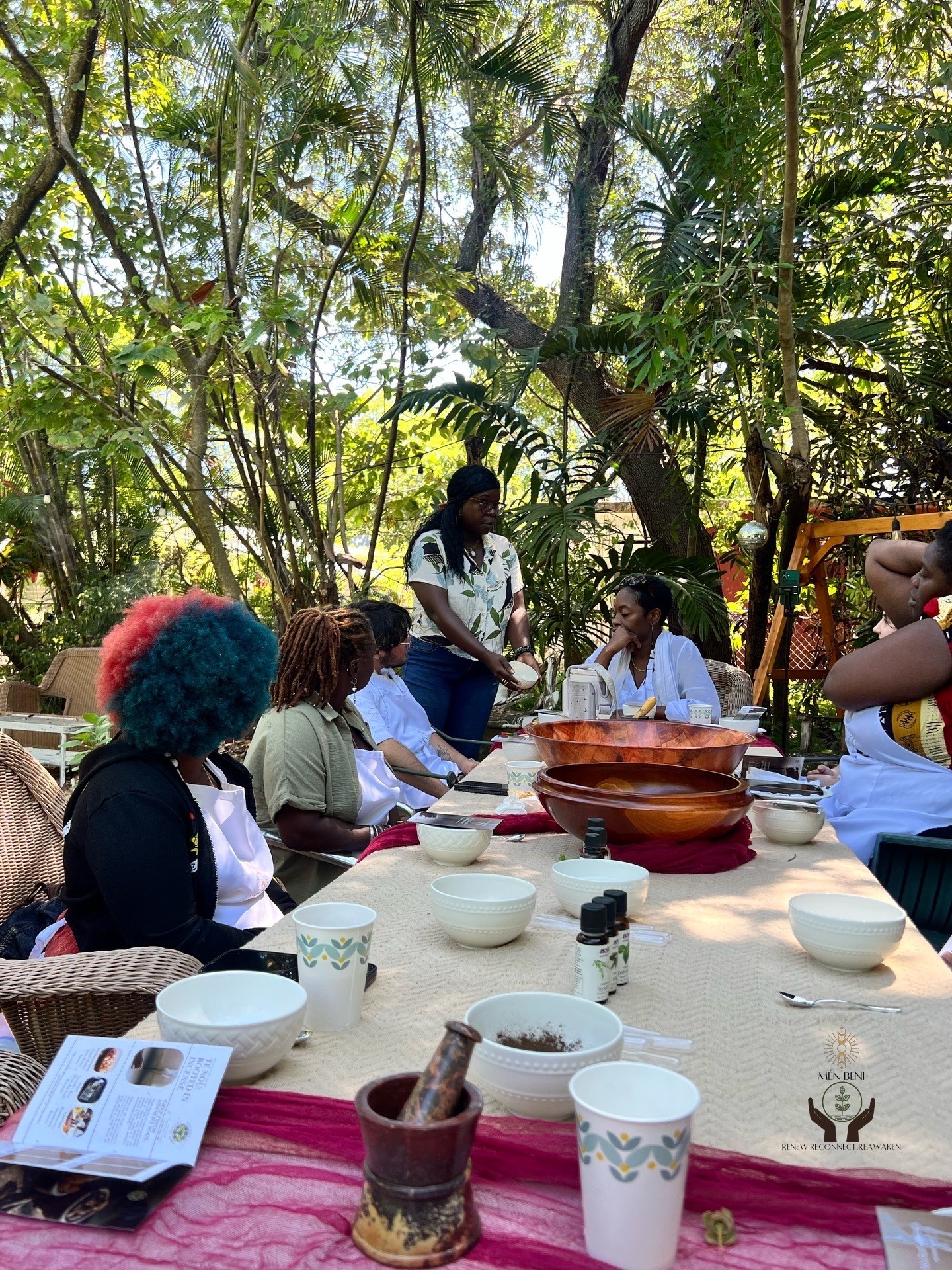 Group of people sitting around a table outdoors with trees and greenery in the background, engaged in a discussion or activity.
