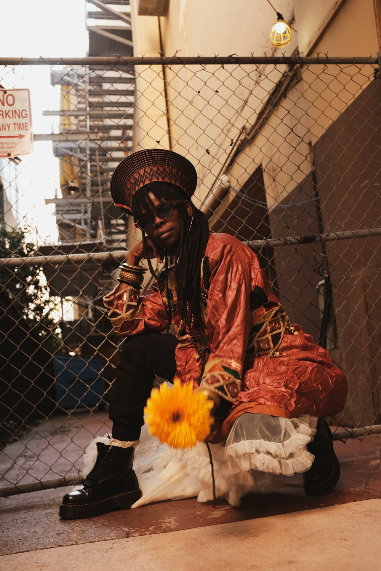 Person wearing a hat, sunglasses, and a red outfit crouches behind a chain-link fence with a yellow flower in front.
