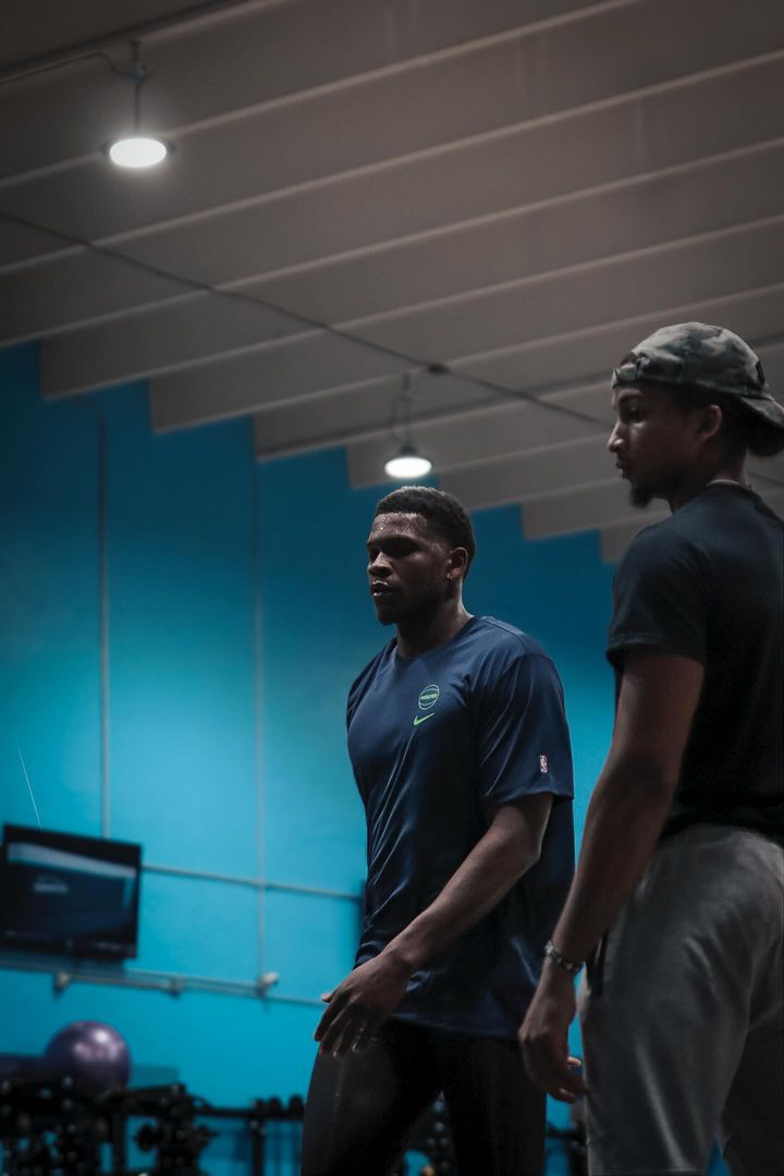 Two men stand in a gym with blue walls and ceiling lights, one wearing a dark shirt and the other in a sports shirt.