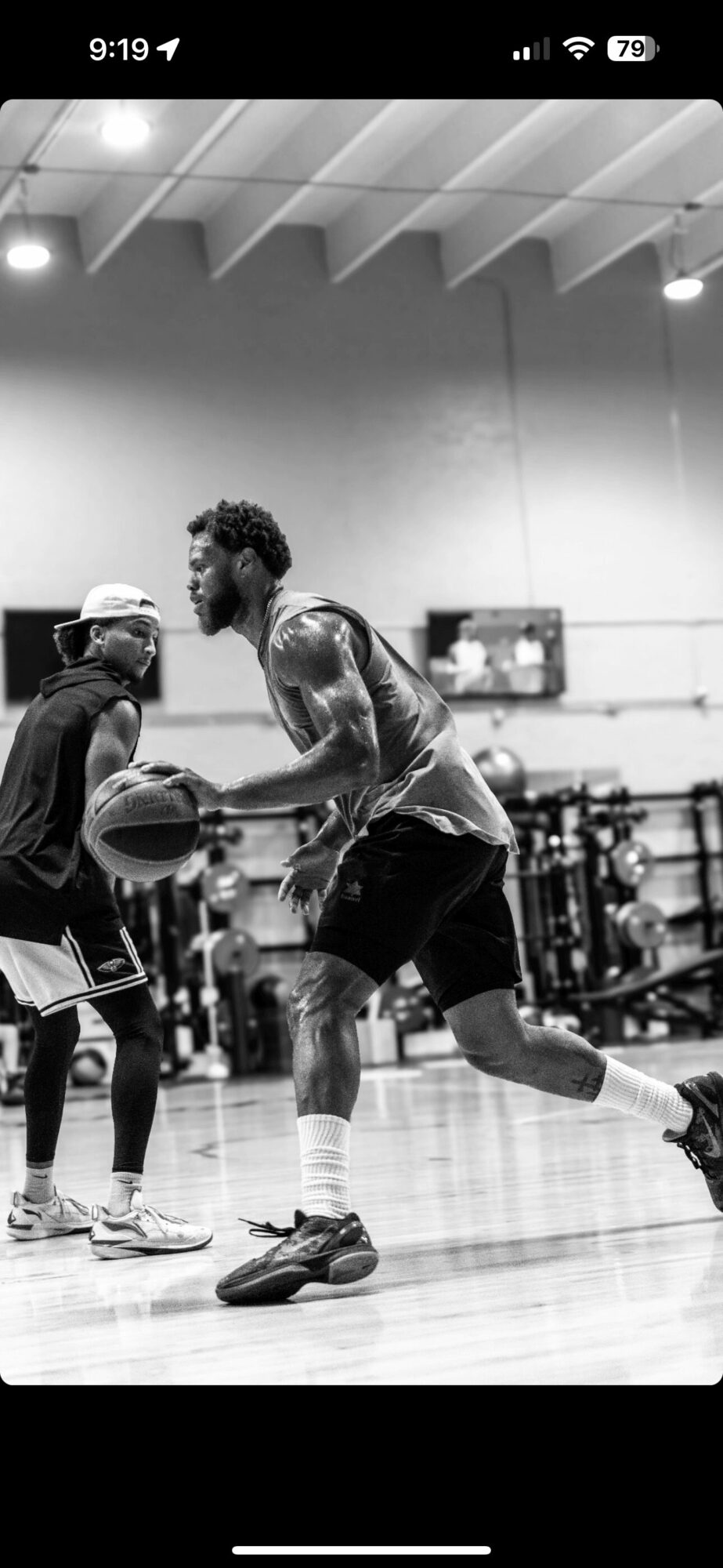 Two basketball players practicing in a gym, one holding a basketball, the other defending, with gym equipment in background.