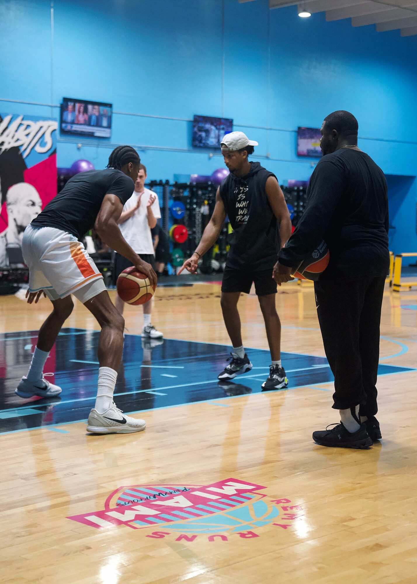 Four people on a basketball court, one holding a basketball, others watching, with blue walls and screens in background.