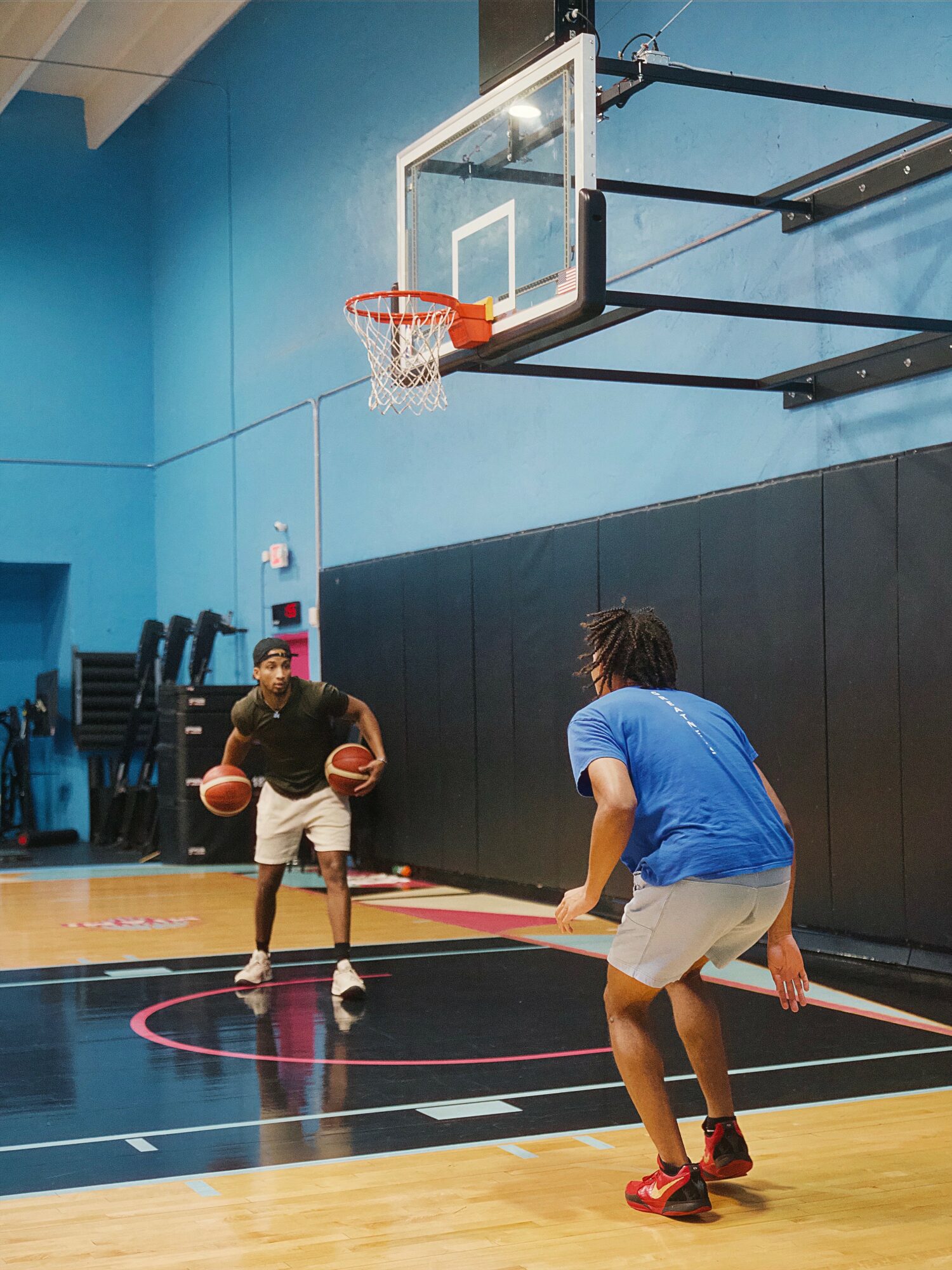 Two people practicing basketball in an indoor gym, one holding a basketball, the other in a defensive stance.
