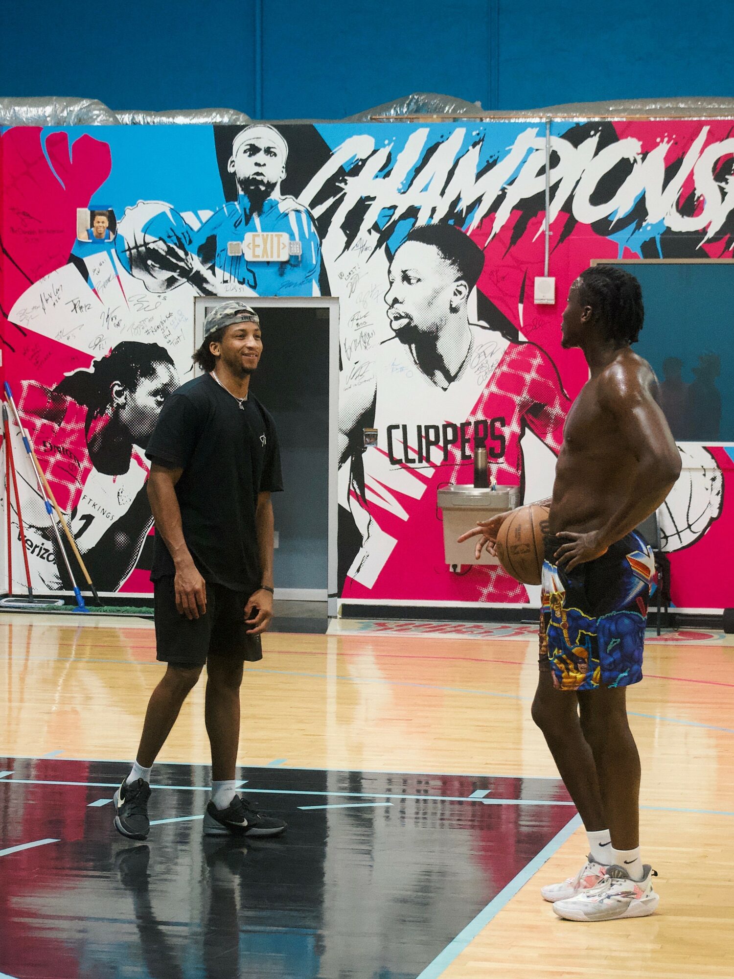 Two men stand on a basketball court with a colorful mural background, one shirtless holding a basketball, the other in black clothing.