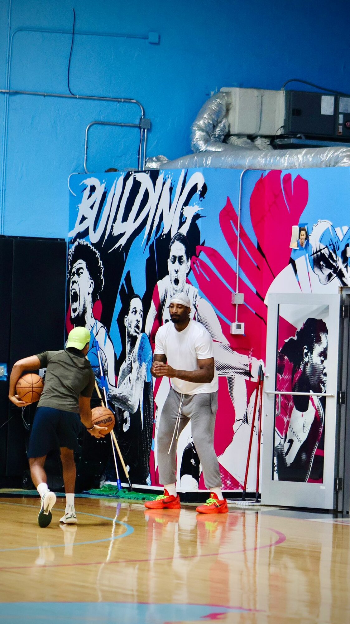 People playing basketball in an indoor gym with colorful wall art and a person taking a photo, gym floor visible.