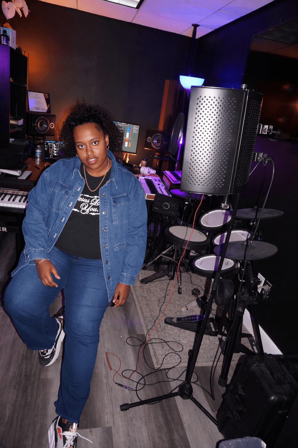 Woman standing in a music studio with equipment, wearing a denim jacket and jeans.