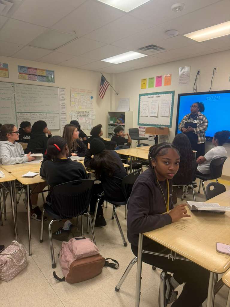Students sit at desks in a classroom, facing a teacher and a large screen, with whiteboards and colorful posters on walls.