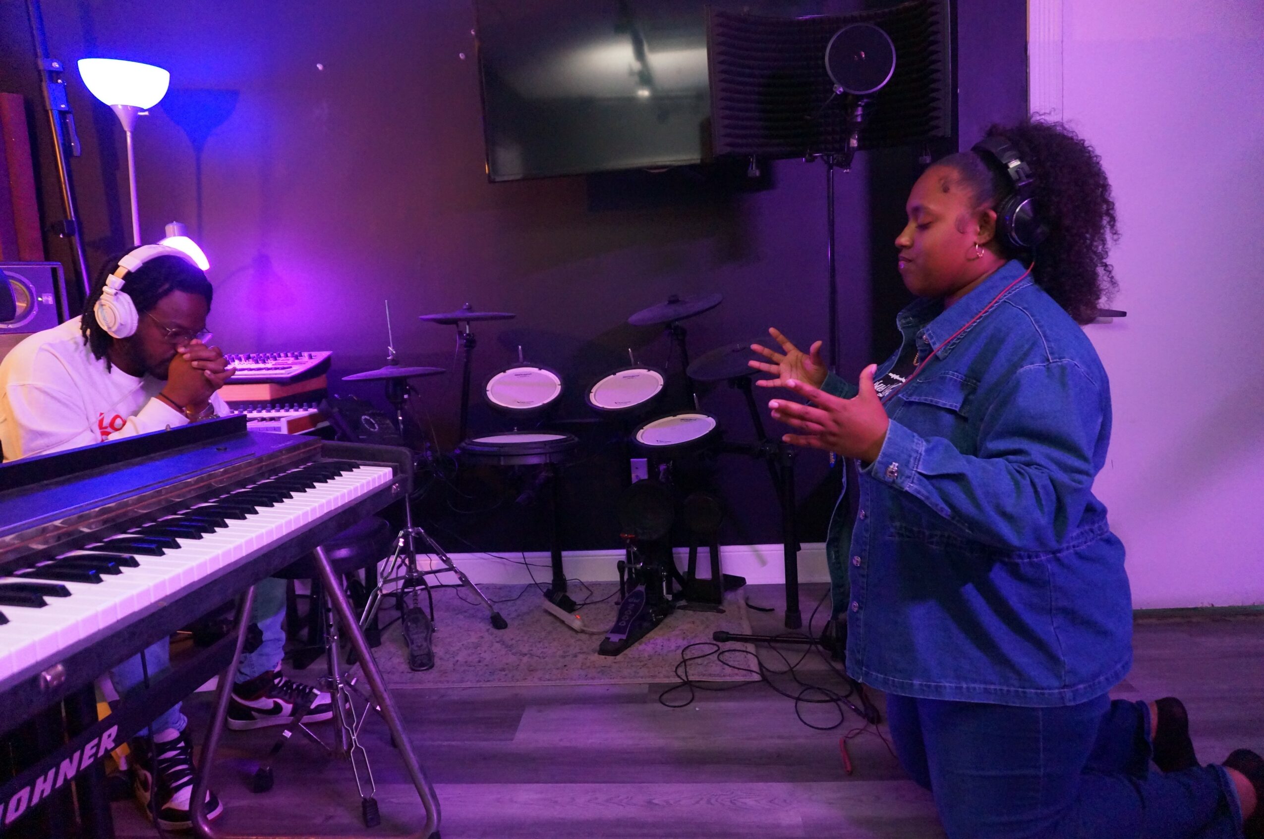 Two women in a music studio, one playing keyboard and the other singing or speaking, with purple lighting and musical equipment.