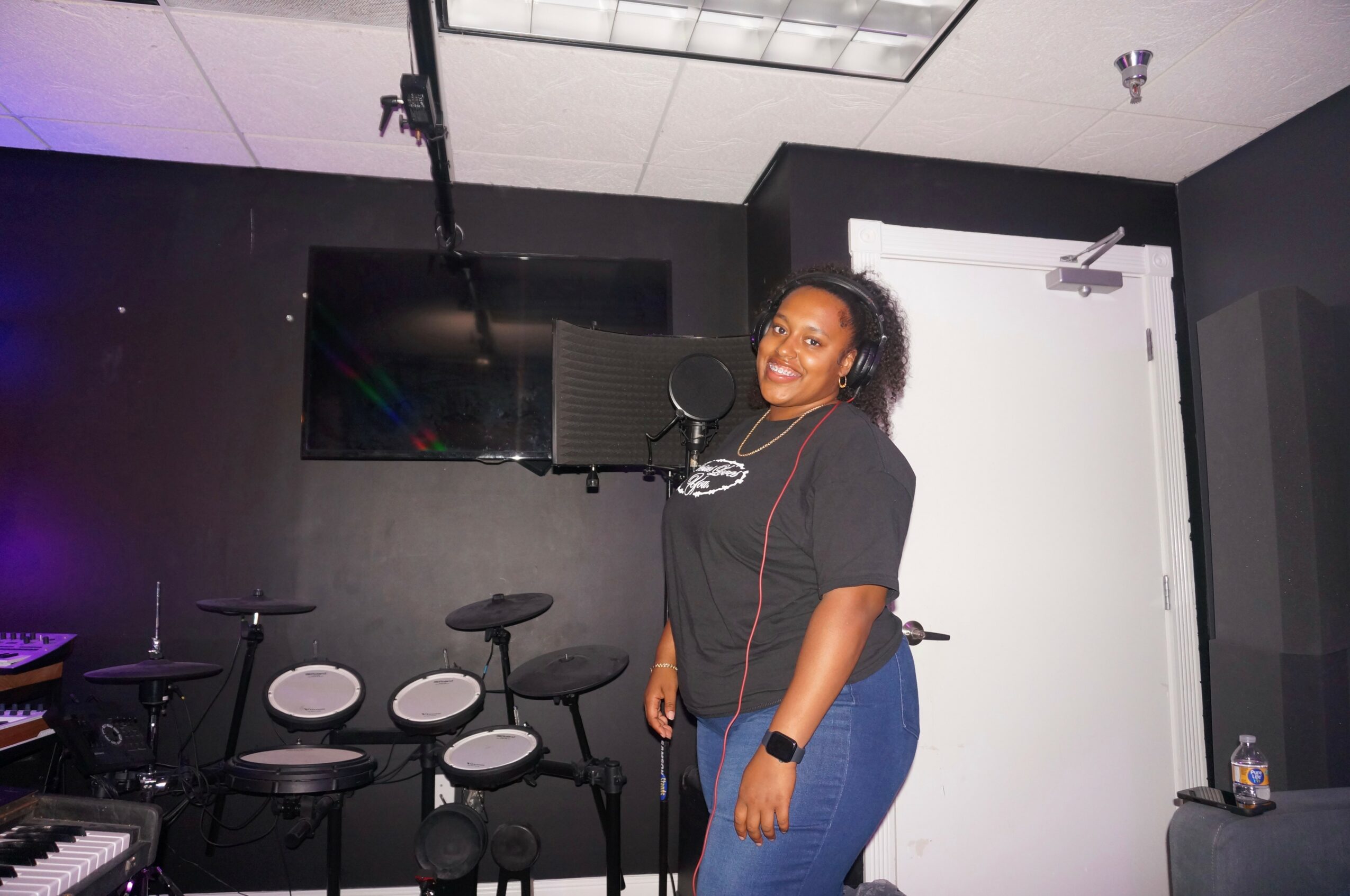 Woman smiling in a recording studio with musical instruments and a large screen behind her.