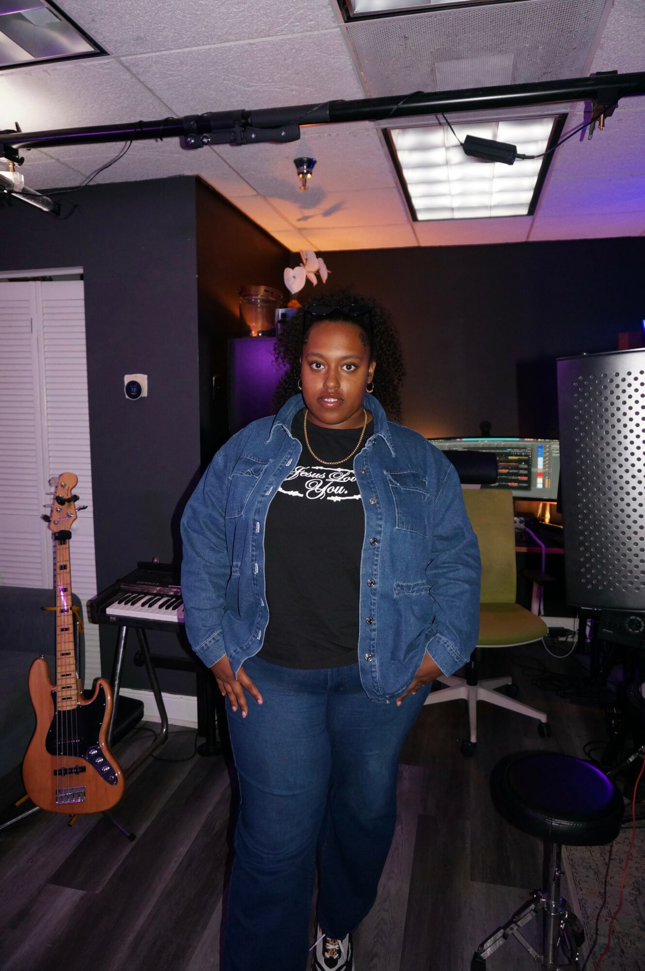 Woman standing in a music studio with a guitar, keyboard, and audio equipment, wearing a denim jacket and black shirt.
