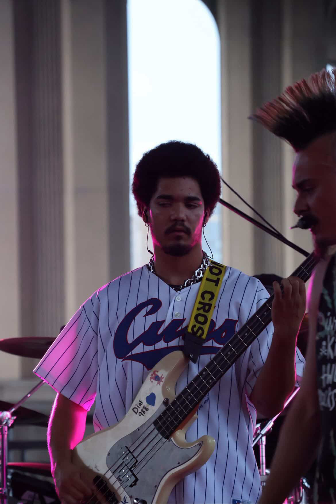 Two musicians perform on stage, one playing a guitar, with a large window in the background.