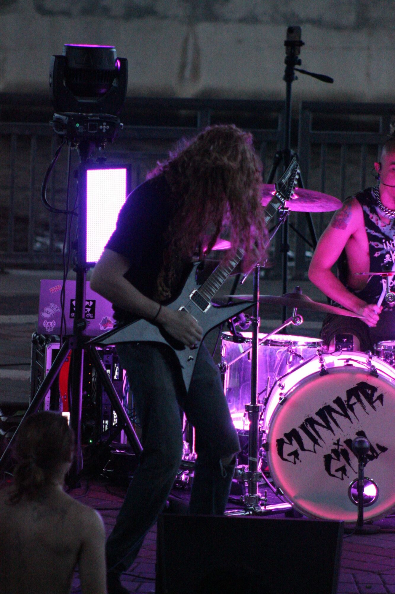 Two women perform on stage with purple lighting, one singing into a microphone and the other playing guitar, with a drum set in the background.