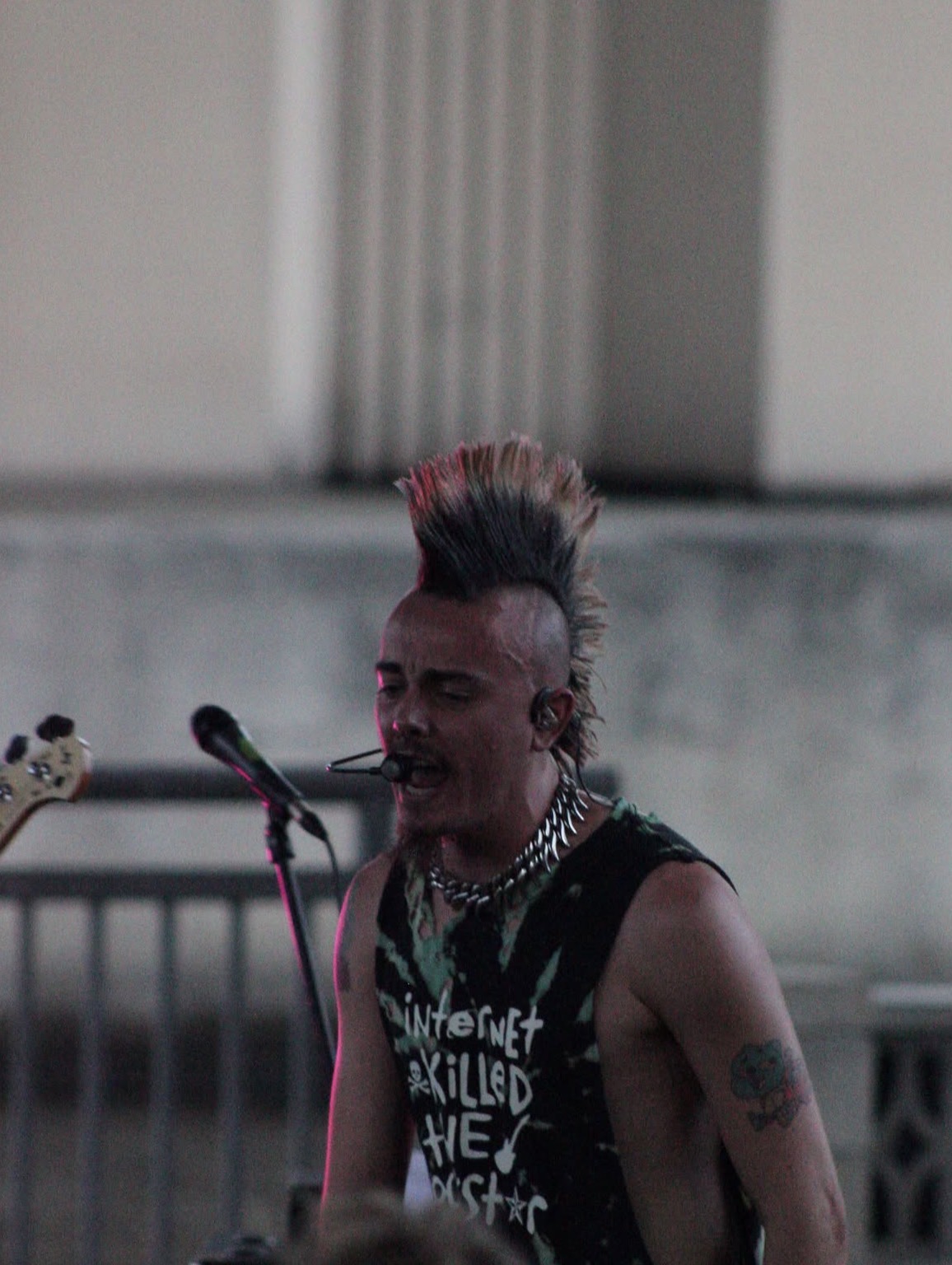 Person with a mohawk hairstyle singing into a microphone outdoors, wearing a sleeveless shirt and jewelry.