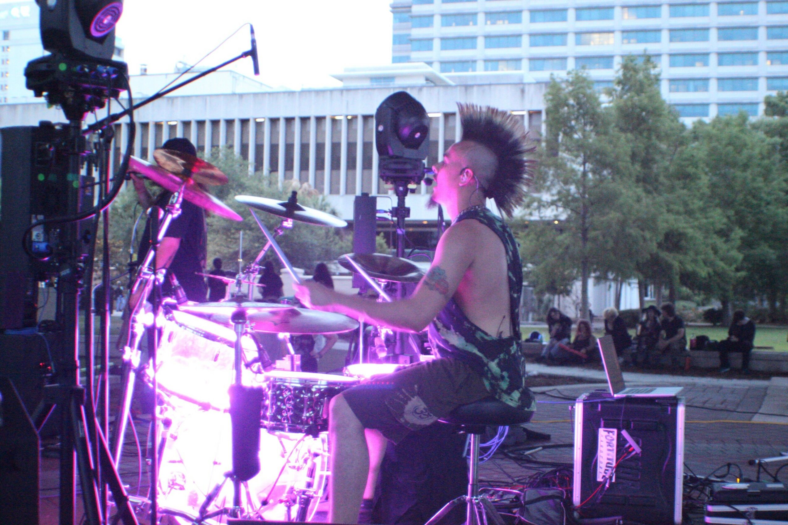 Person playing drums outdoors with purple lighting, surrounded by equipment and trees, in an urban setting.