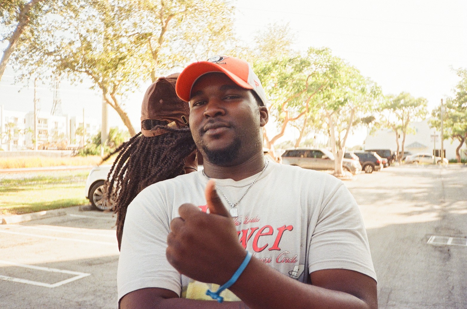 Young man wearing a red cap and white T-shirt giving a thumbs-up outdoors, with trees and parked cars in background.