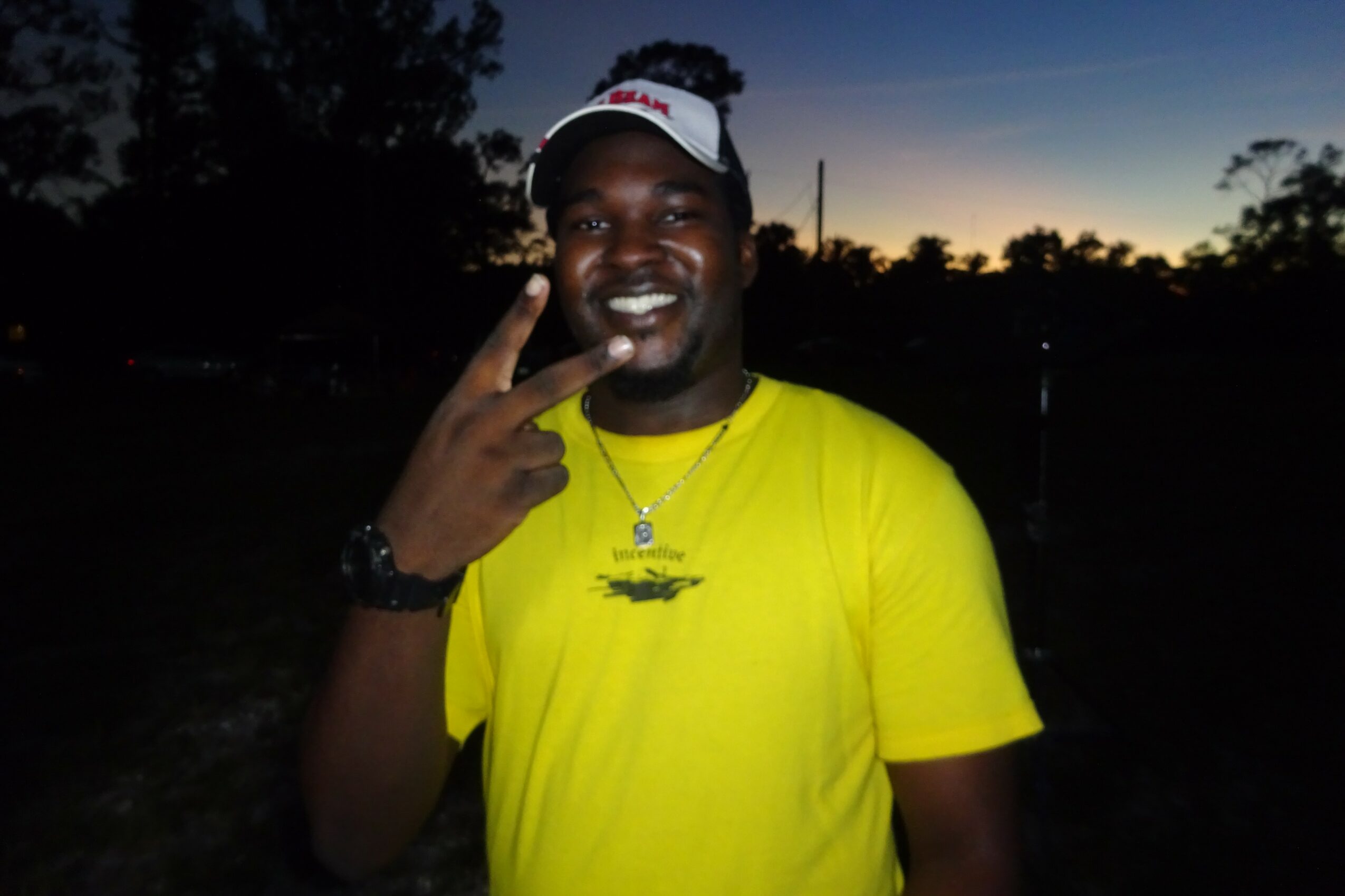 Person smiling outdoors at sunset, wearing a yellow shirt and a white cap, making a peace sign with hand.