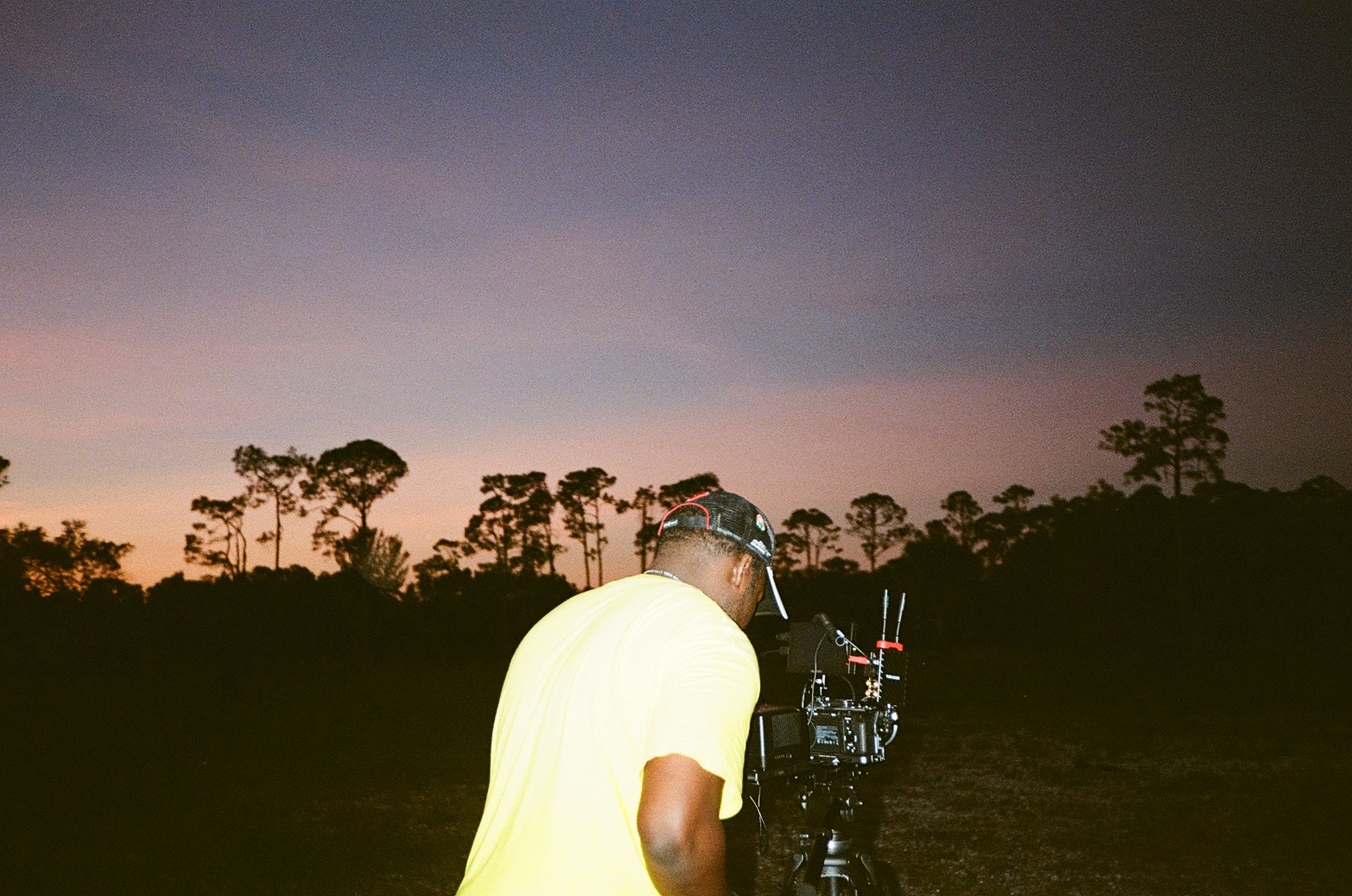 Person in yellow shirt operating a camera outdoors during sunset or dusk, with trees in the background.