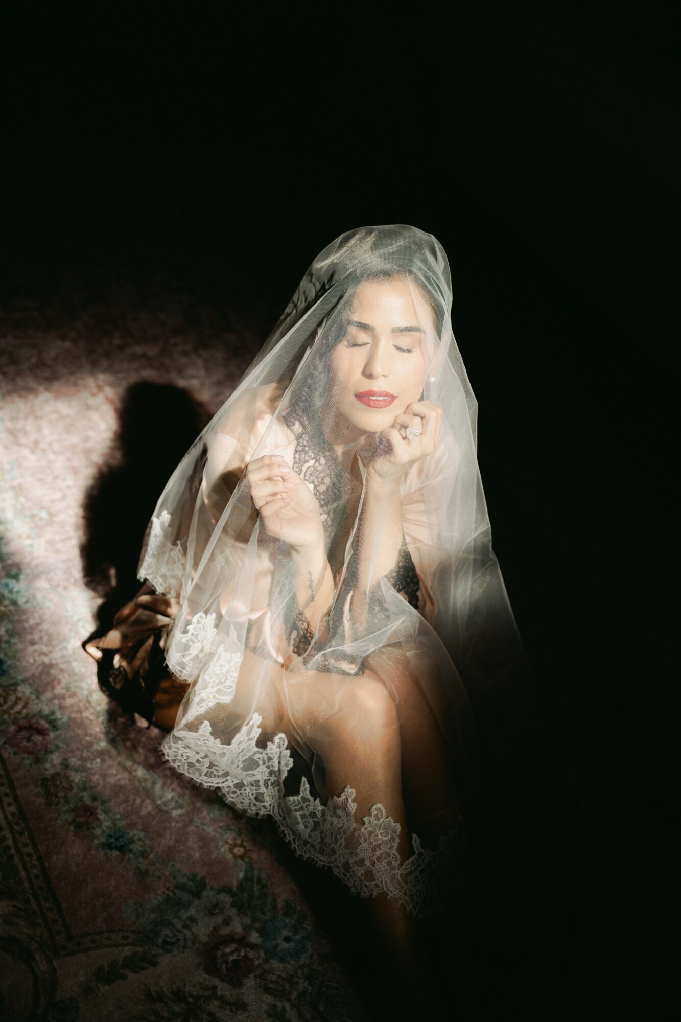 Woman with closed eyes, wearing a veil, sitting on a dark background, illuminated softly, with lace details on her dress.