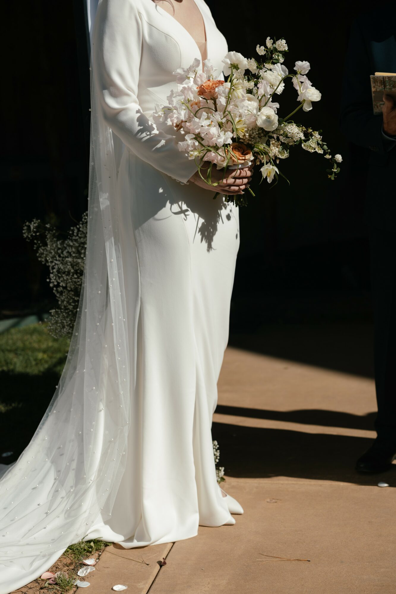 Person in white wedding dress holding a large bouquet of white and pink flowers, standing outdoors.