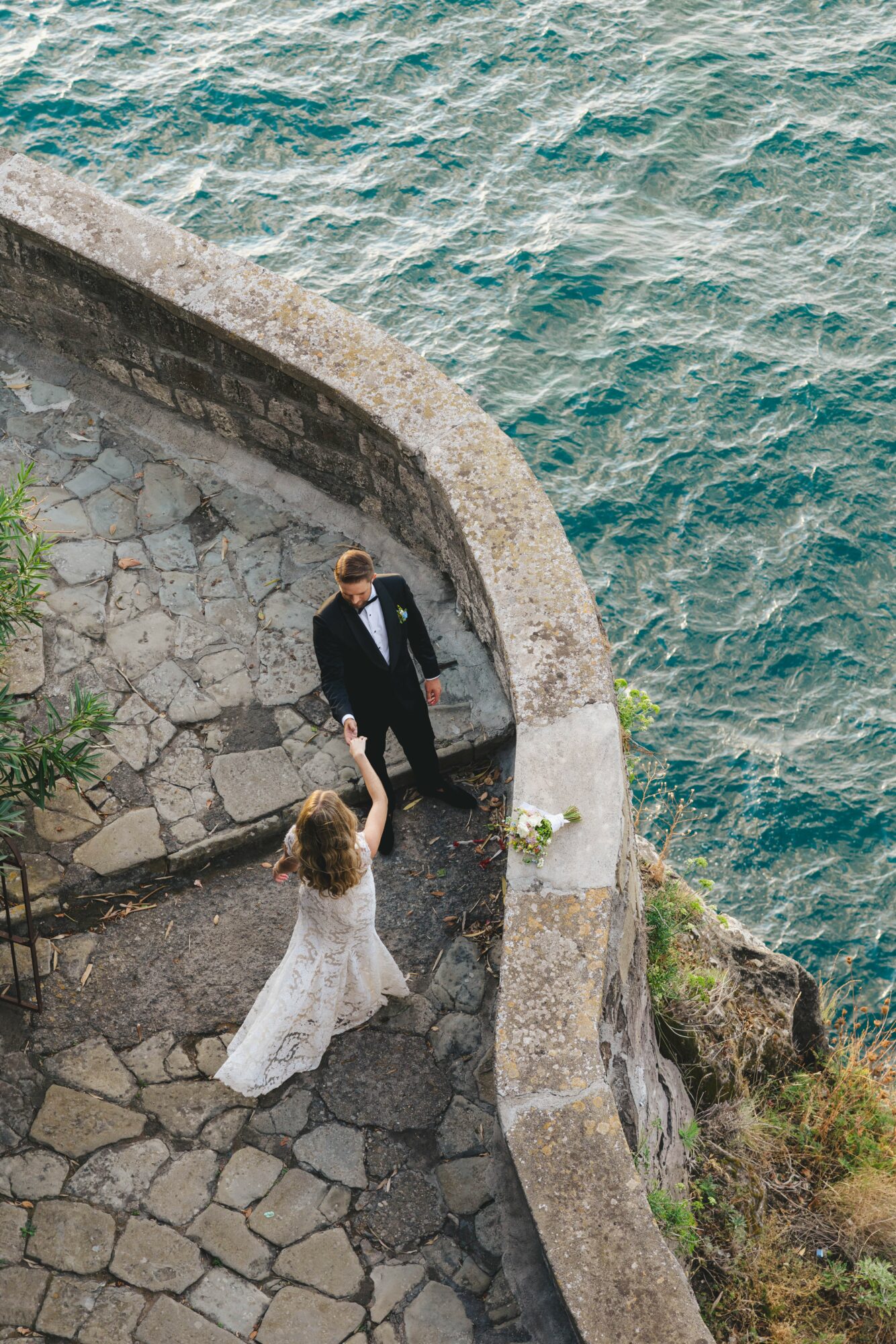 A couple in wedding attire stands on a stone pathway by the water, with a curved stone wall behind them.
