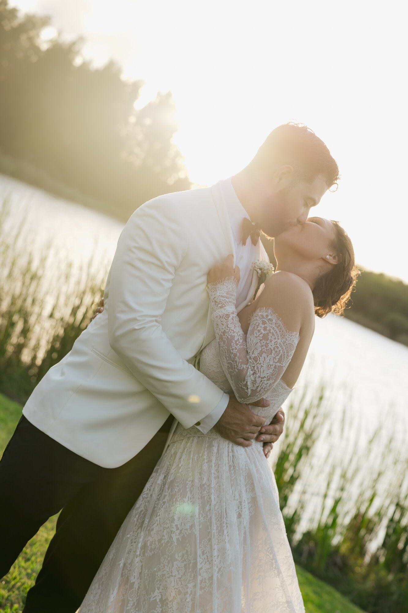 A couple in wedding attire sharing a kiss outdoors near a body of water, with sunlight behind them.