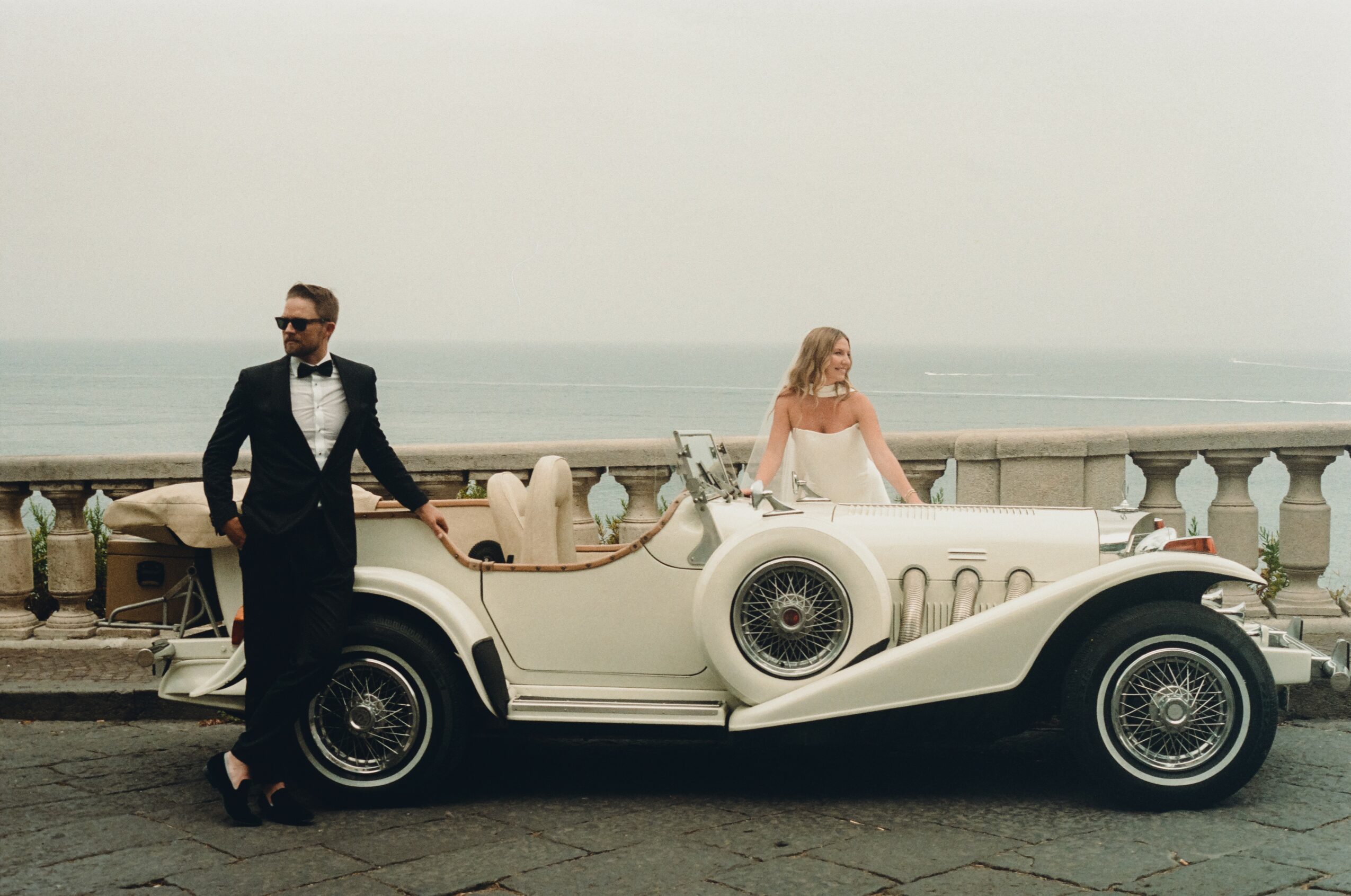 Man in tuxedo and woman in dress stand beside vintage convertible car on seaside promenade.