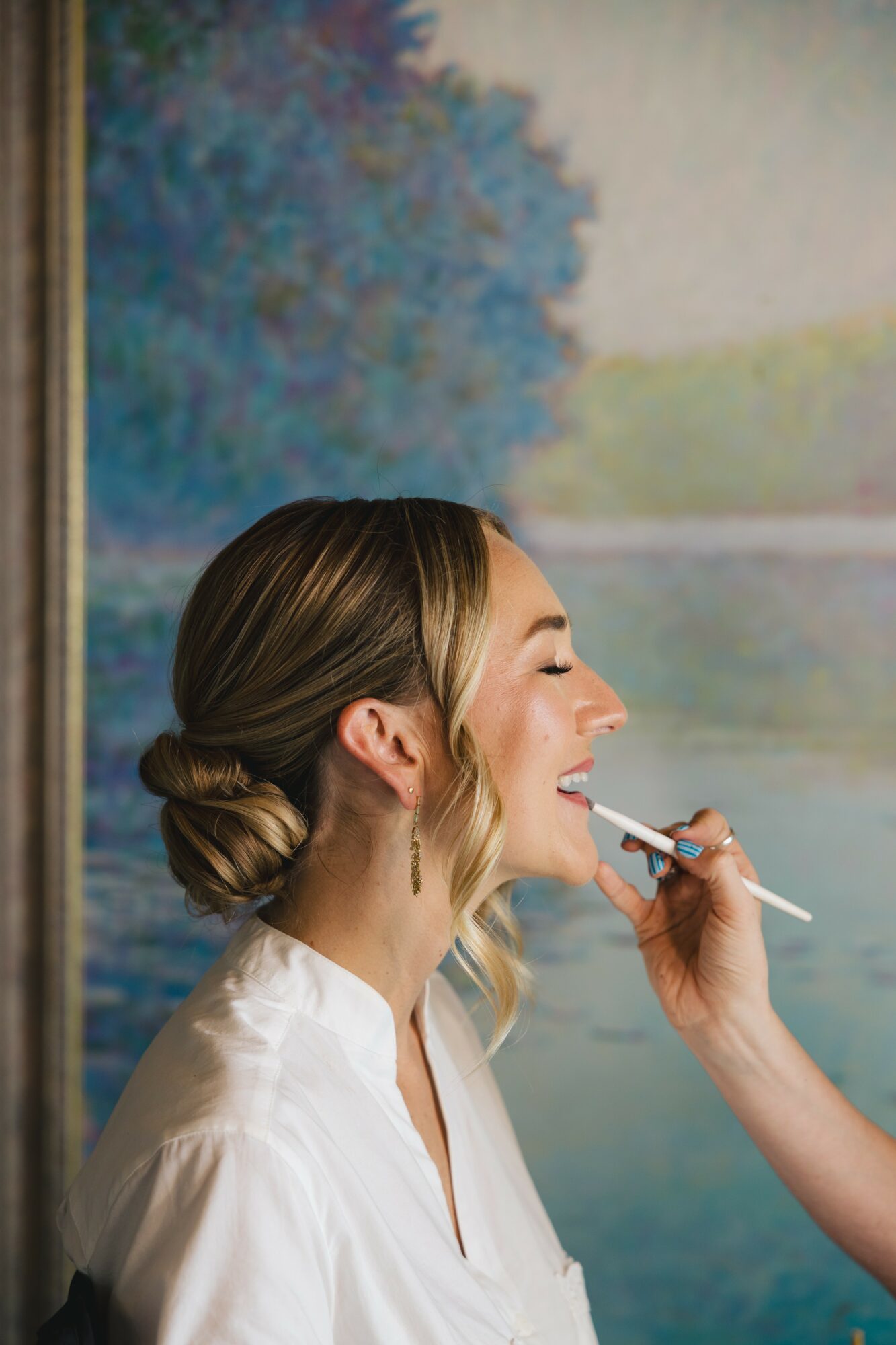 Woman with styled hair getting makeup applied, outdoor background with trees and water, side profile, smiling, wearing white shirt.