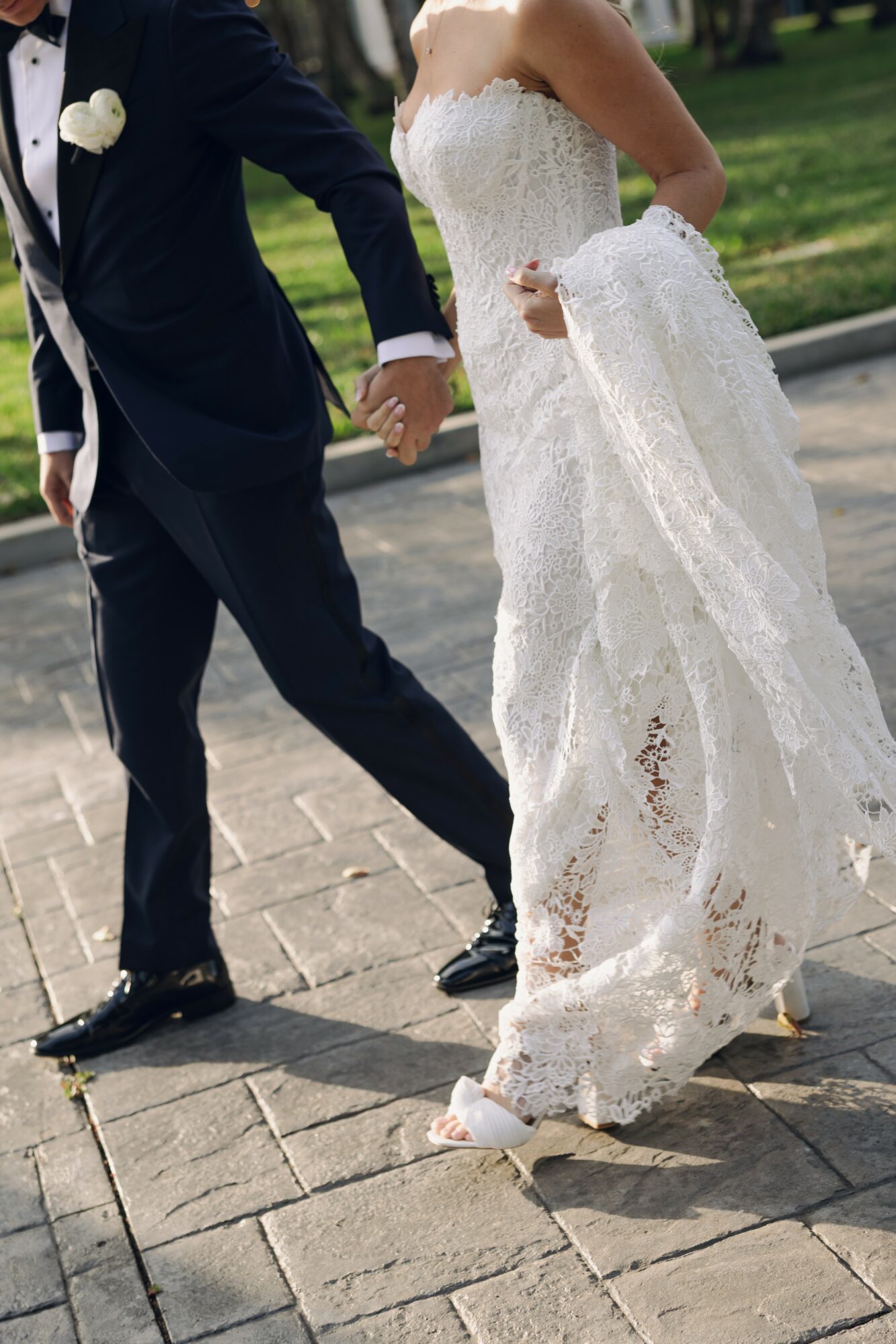 Couple holding hands, walking outdoors; woman in a white lace wedding dress, man in a dark suit, on paved path.