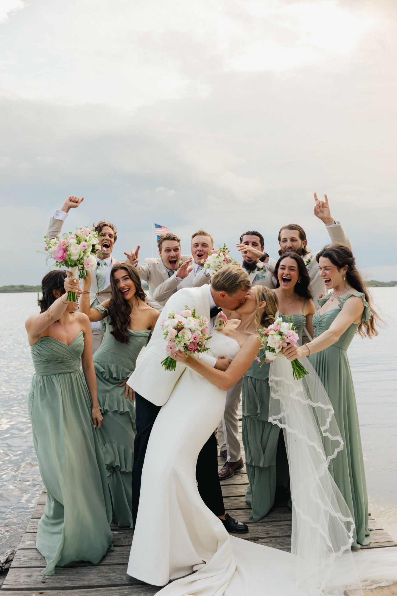Bride and groom kissing on a dock with wedding party celebrating around them, holding bouquets, over water and cloudy sky