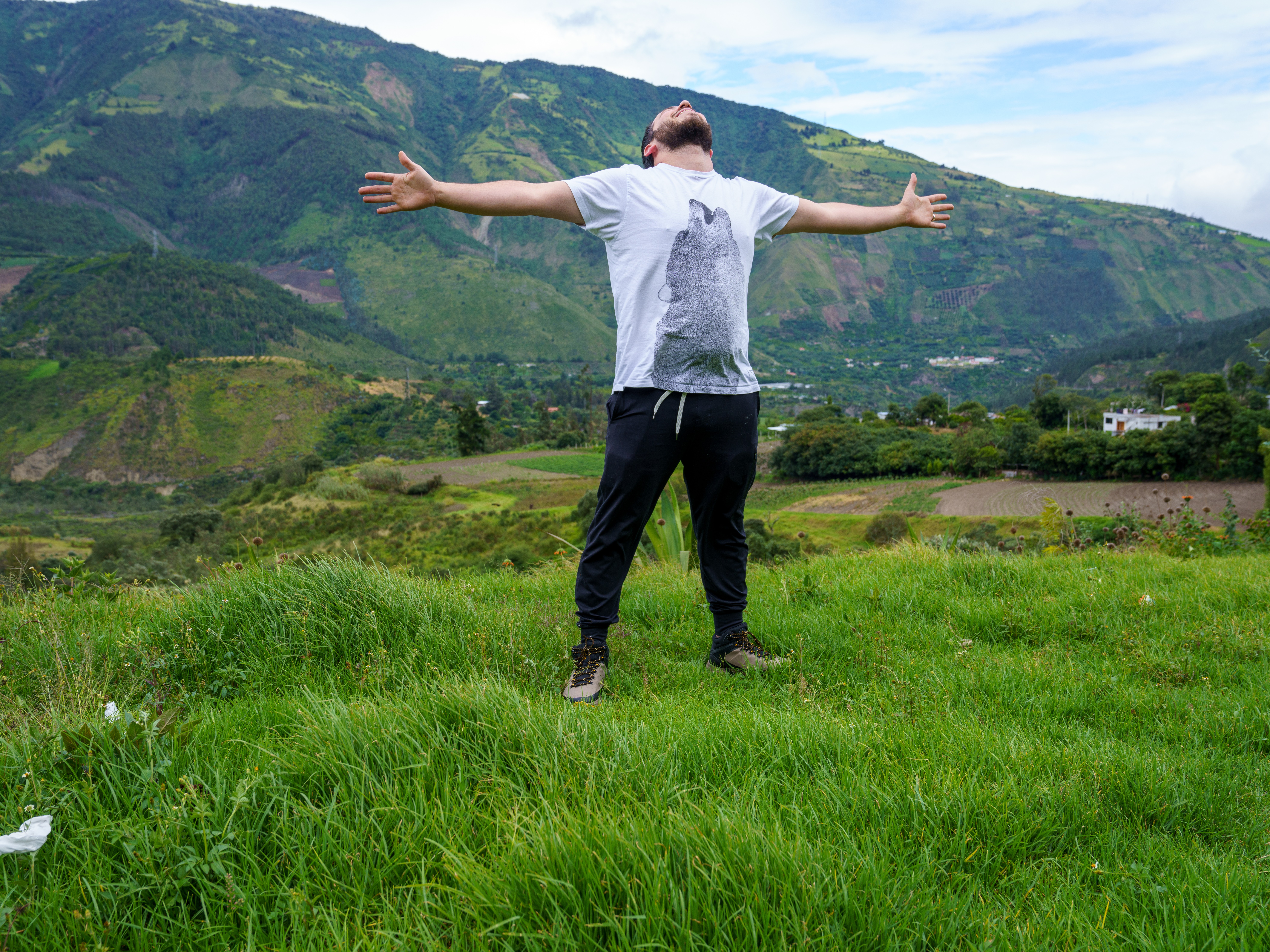 Person standing on grassy field with arms outstretched, mountains and sky in background.