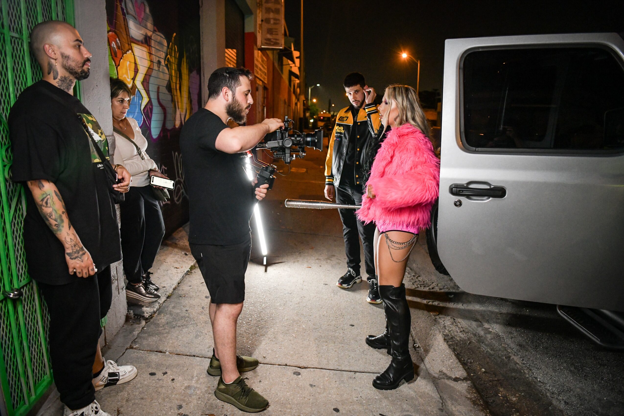 Group of people on sidewalk at night, woman in pink fur coat and thigh-high boots, man with camera, others nearby, city street scene.