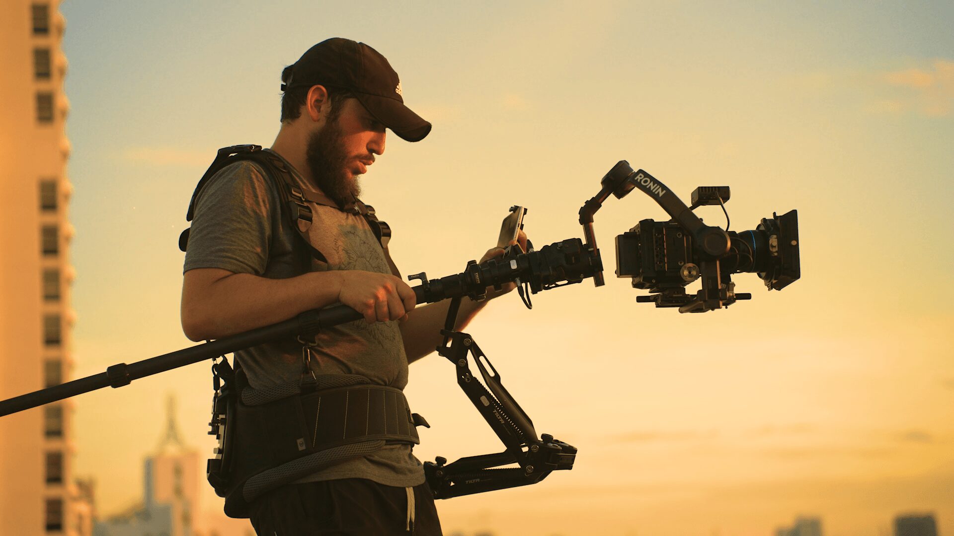 Person operating a camera rig outdoors during sunset, wearing a cap and backpack, with a cityscape in the background.