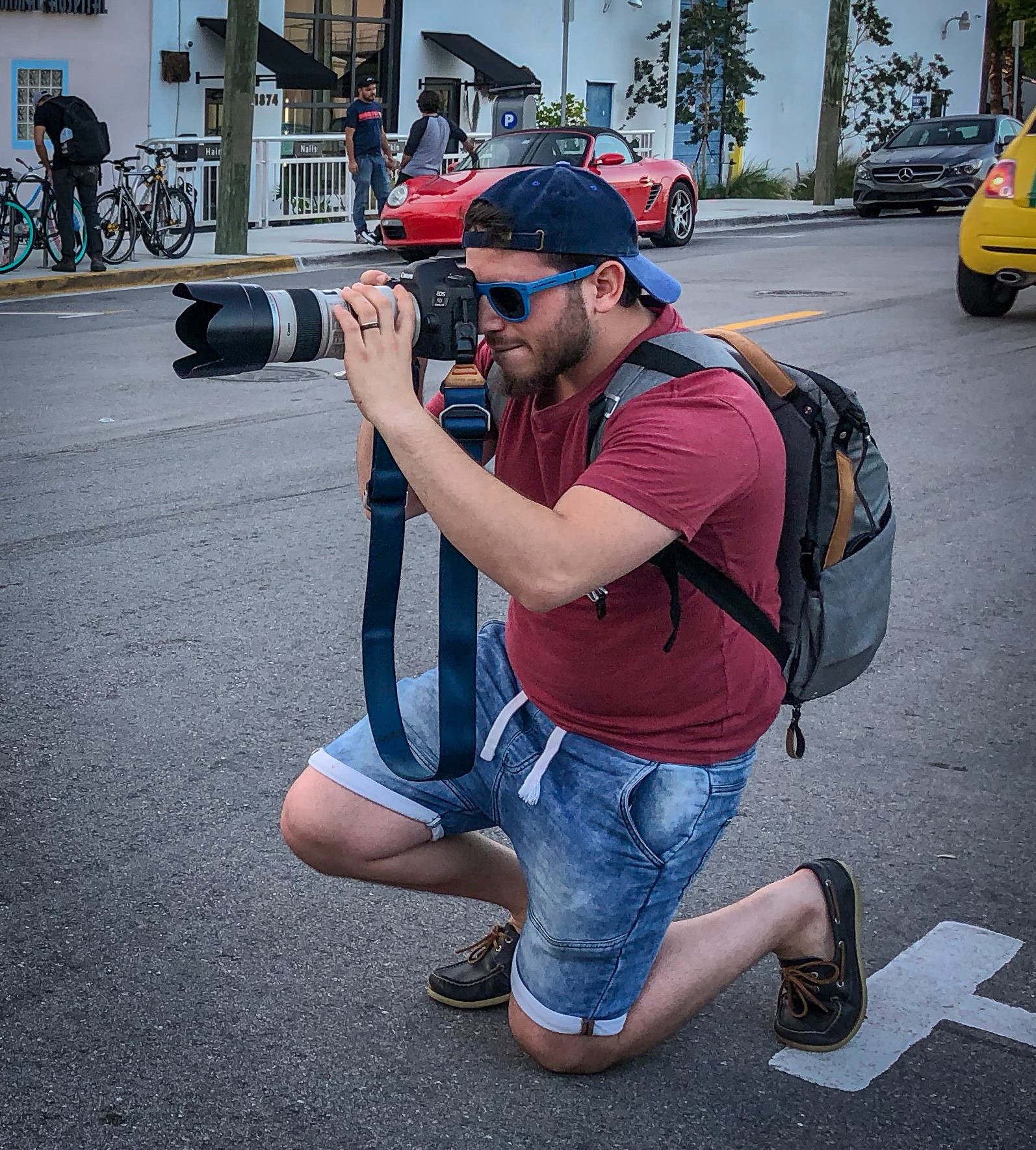 Man kneeling on street with camera, wearing sunglasses, cap, red shirt, denim shorts, backpack, taking photos.