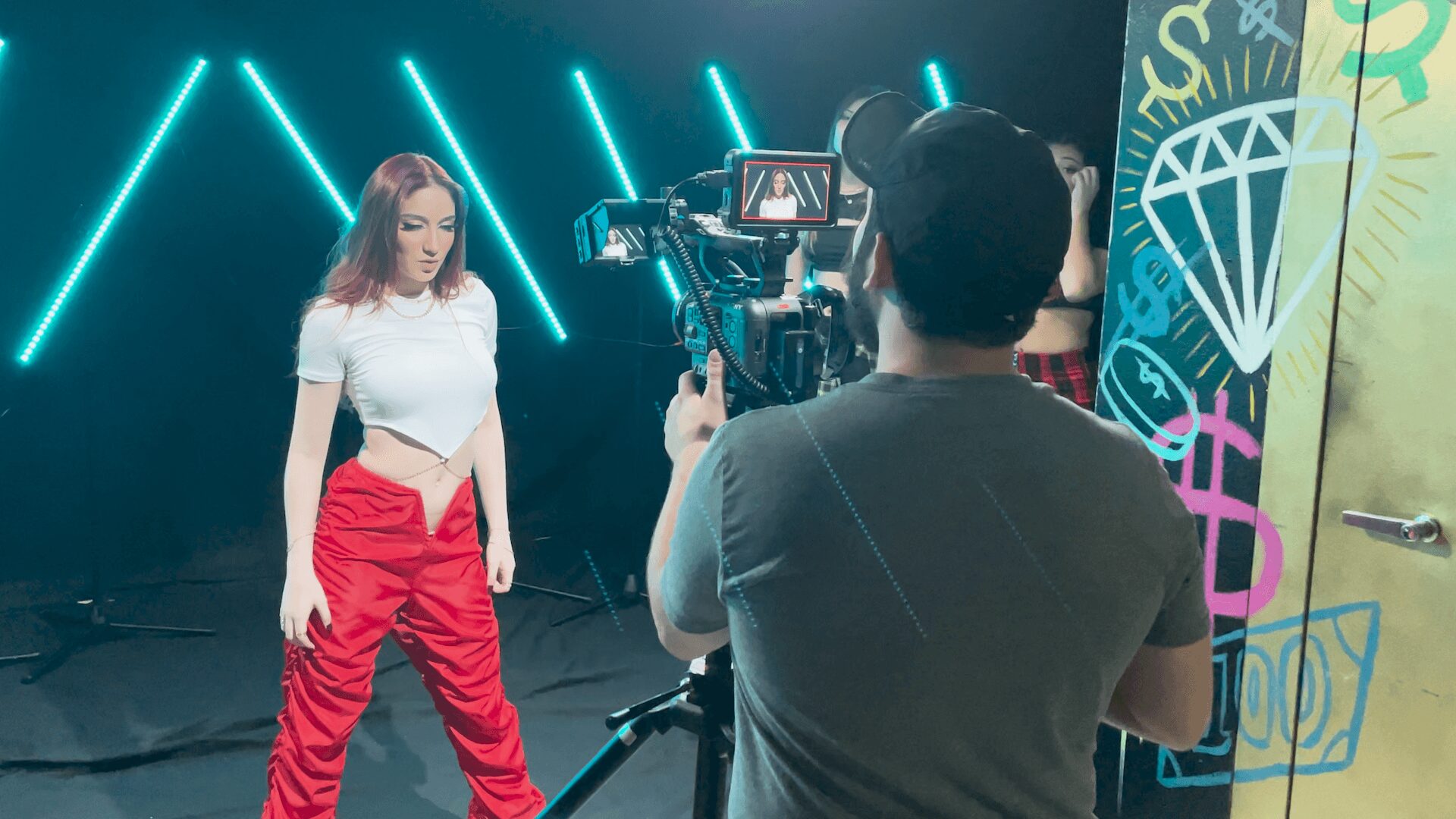 A woman stands on a stage with neon lights, being filmed by a cameraman, with colorful graffiti on the wall nearby.
