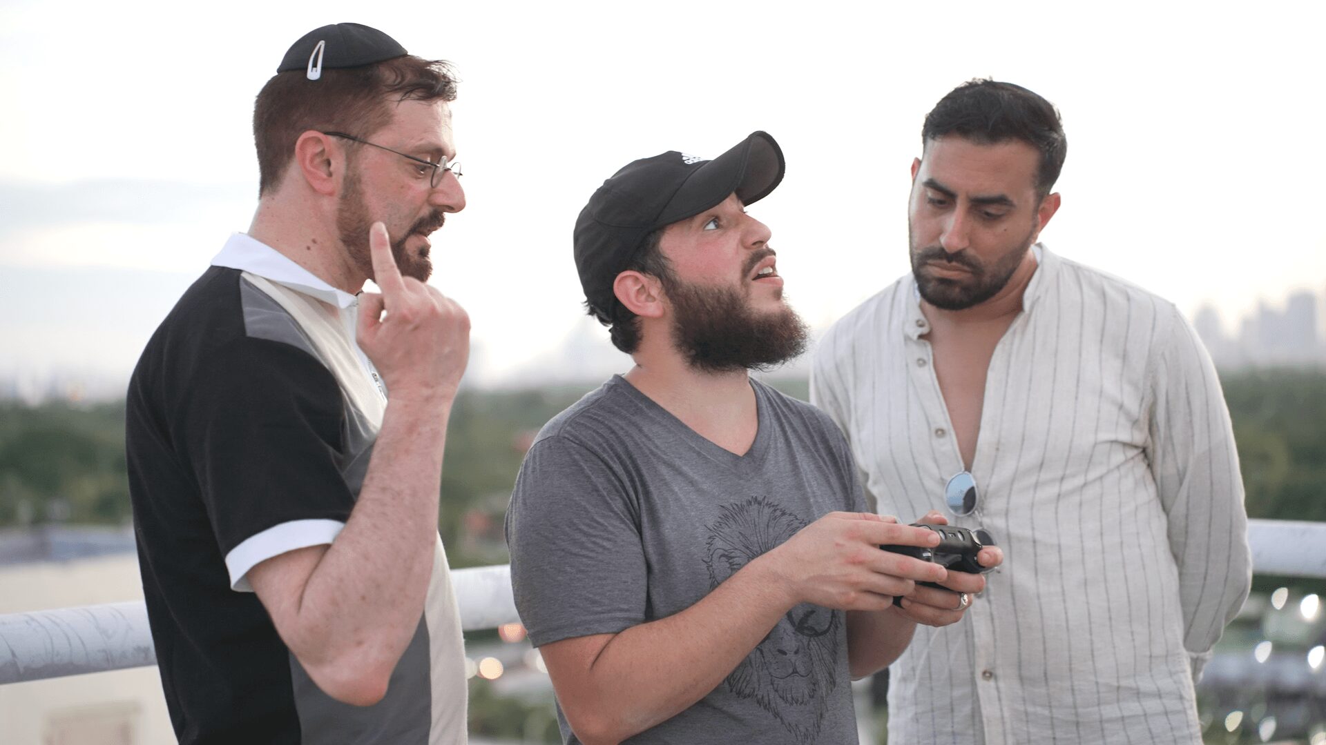 Three men standing outdoors, one showing something on a phone to the others, with a blurred background.