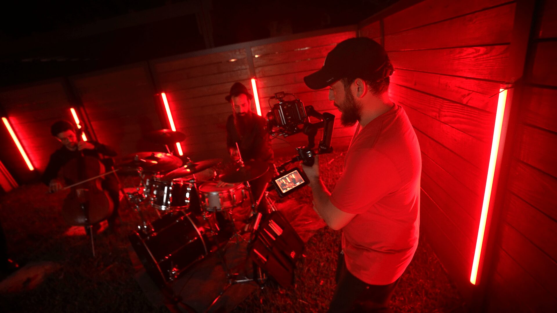 Person operating camera equipment in a dimly lit room with red vertical lights, a drum set, and other musicians in the background.