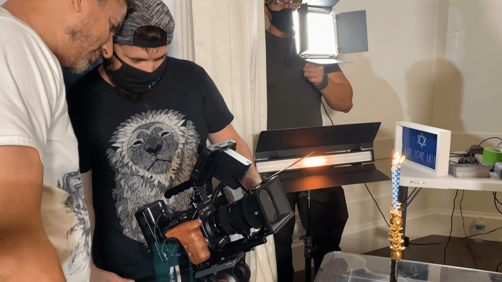 Three people in a room, one holding a camera, with a lit candle on a table, and a computer monitor in the background.