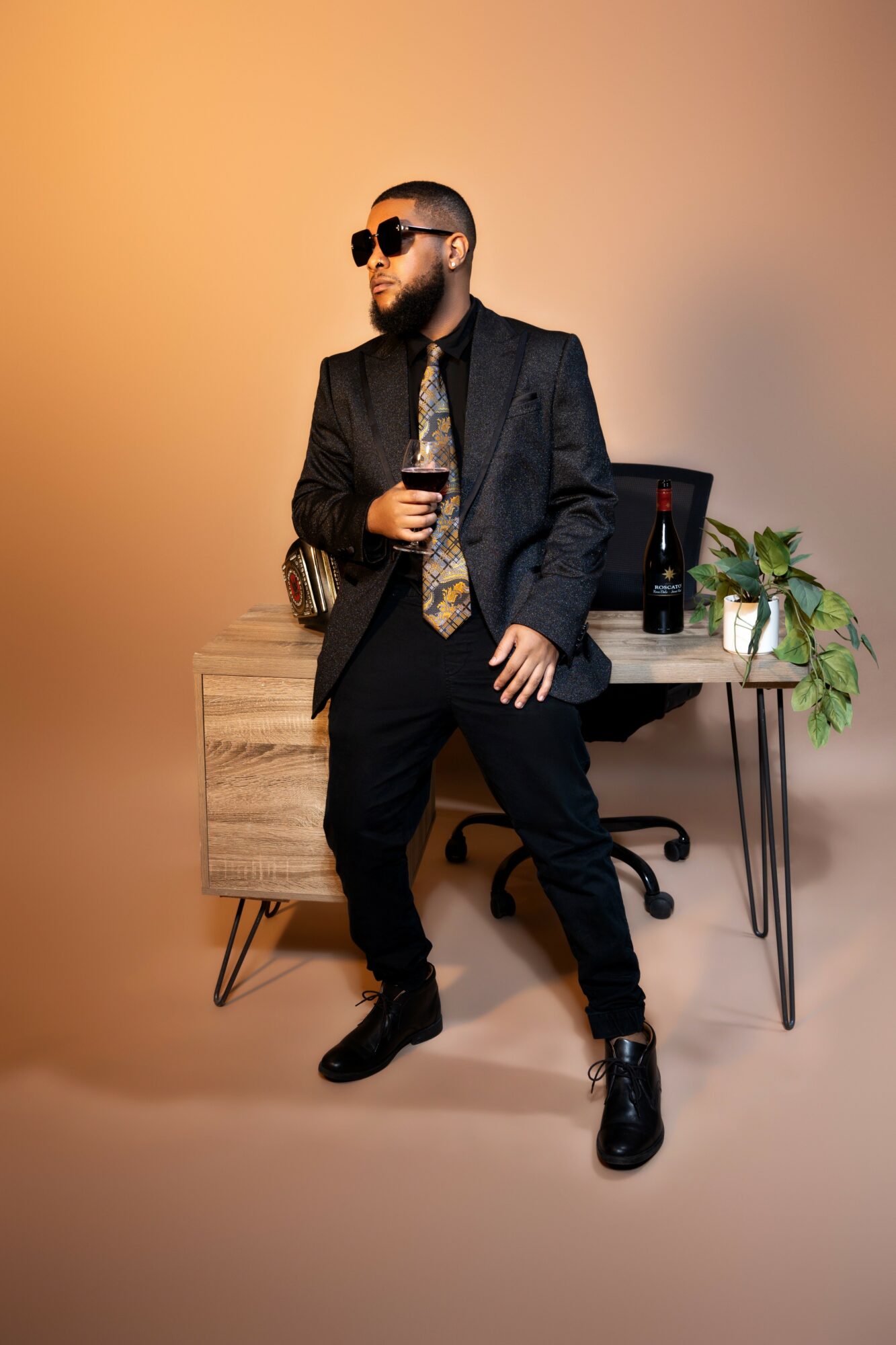 Man with sunglasses and beard in a suit holding a glass, standing in front of a desk with a plant.