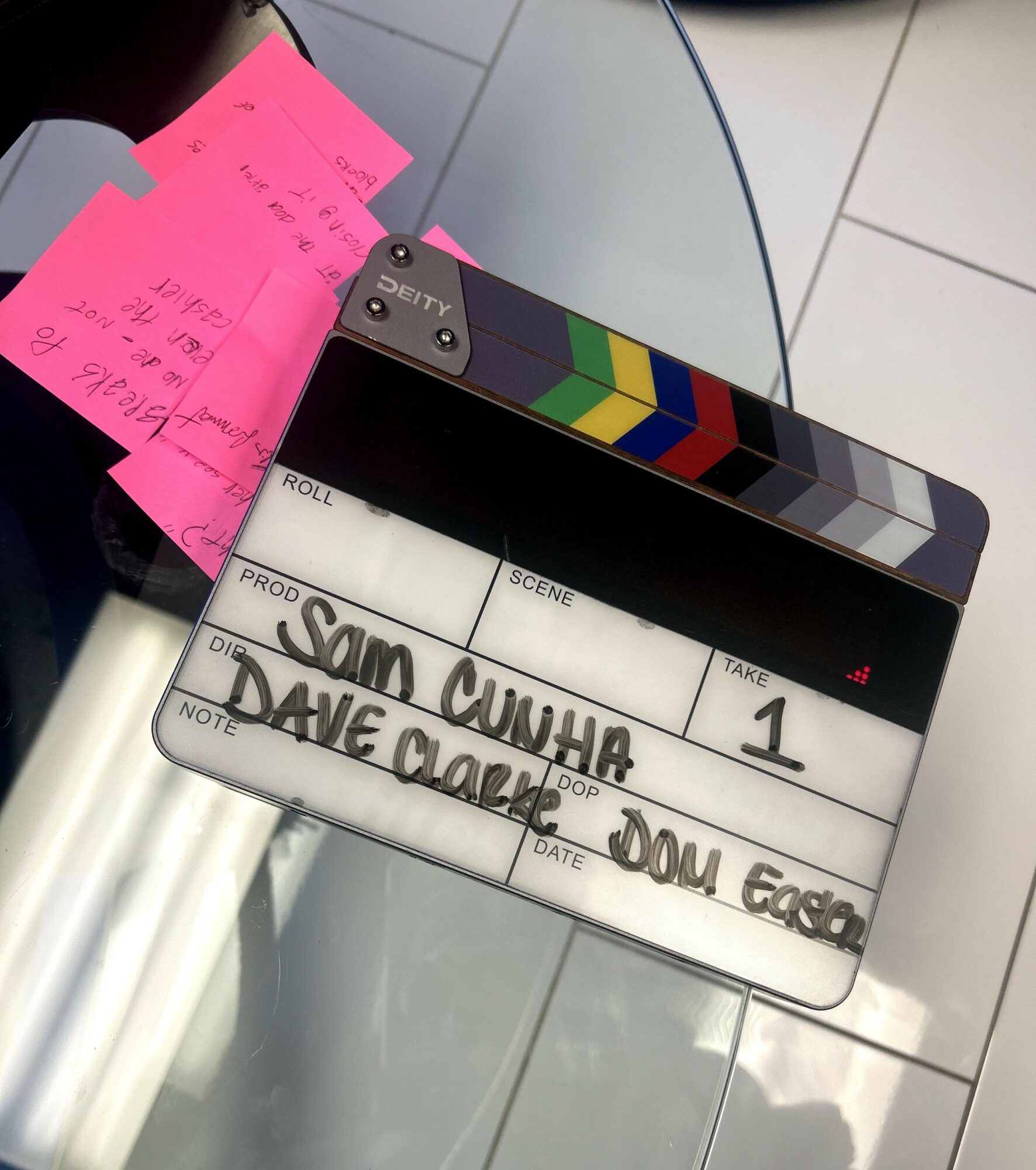 Clapperboard with handwritten text, on a glass table, with pink notes nearby, tiled floor in background.
