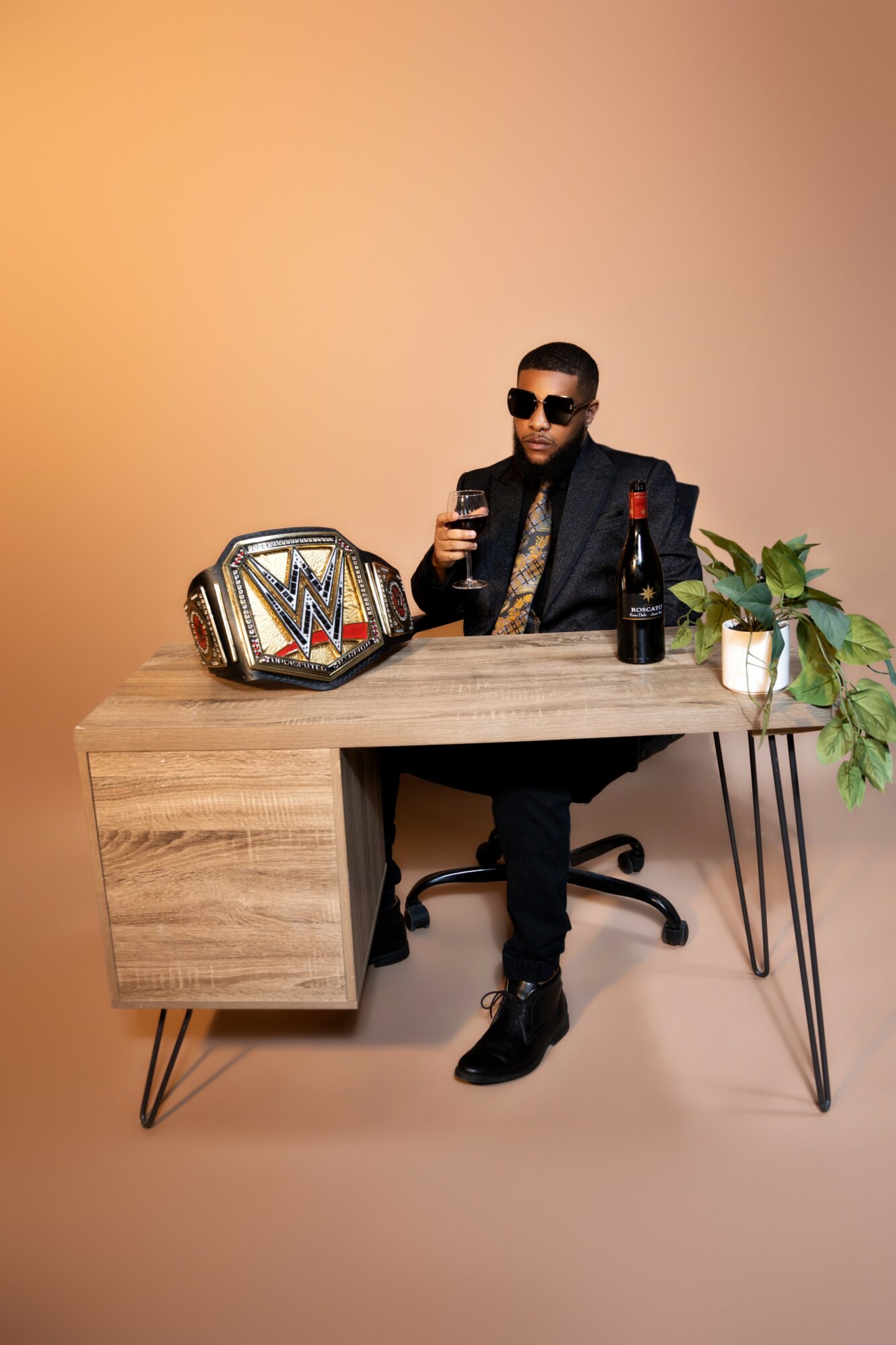 Man sitting at a desk with a championship belt, a bottle, and a plant, wearing sunglasses and a suit, smiling.