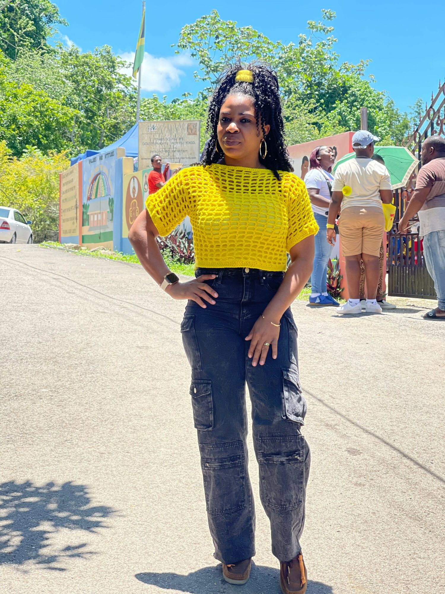 Woman in yellow top and black pants standing outdoors with trees and people in background.