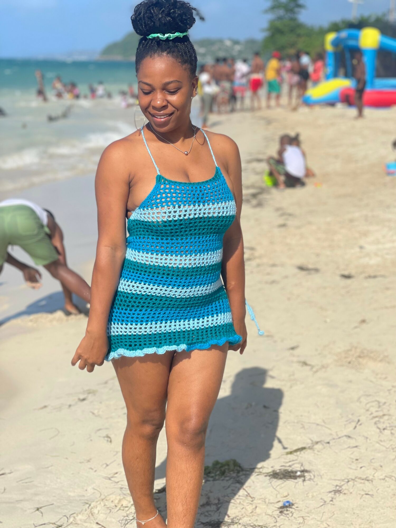 Smiling woman in a blue striped swimsuit walking on sandy beach with people and water in background.