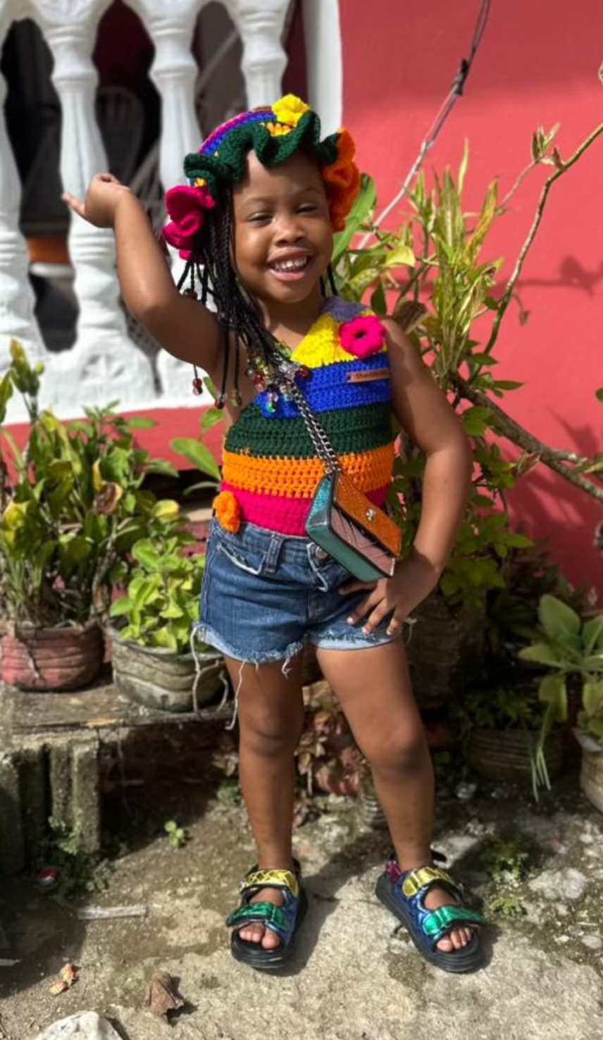 Young girl smiling outdoors, wearing a colorful striped top, denim shorts, and sandals, surrounded by plants and a pink wall.