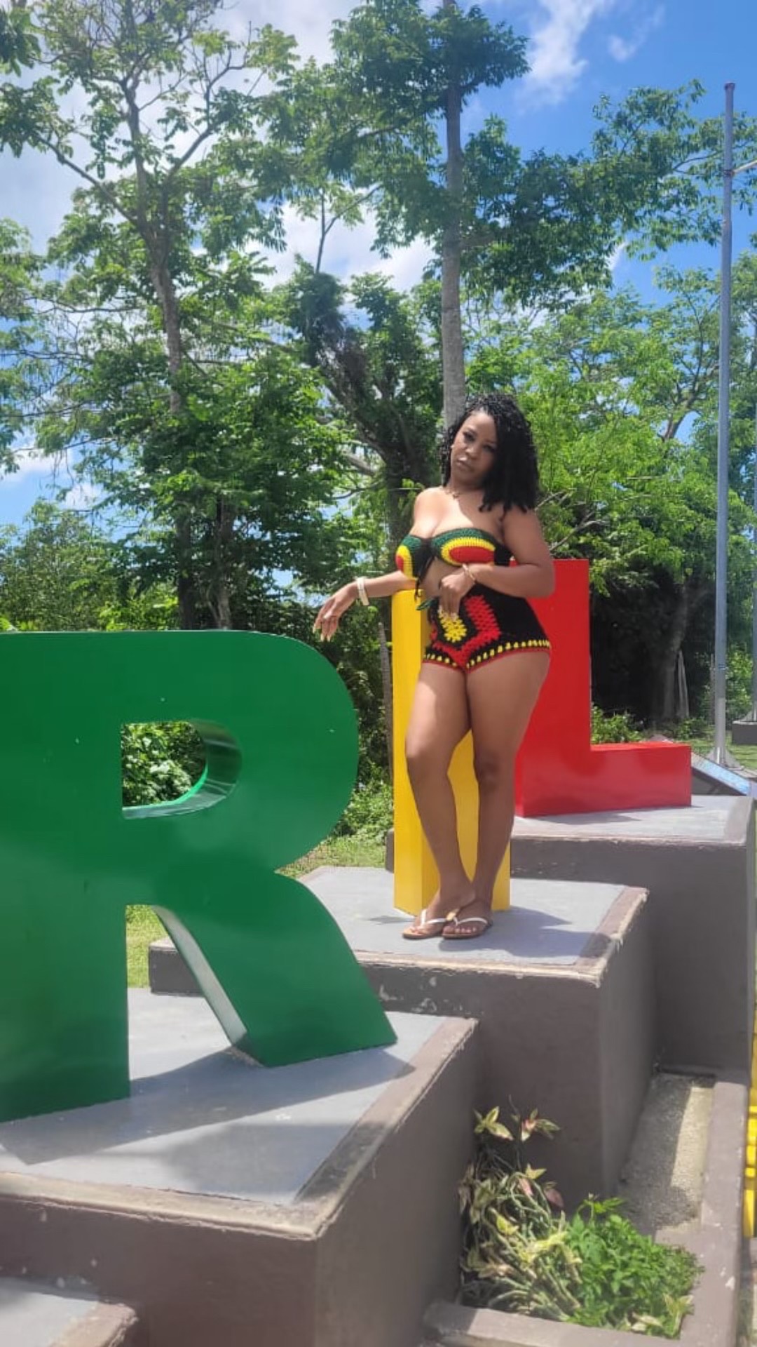 Woman in colorful outfit standing on a concrete platform with large colorful letters, trees and blue sky in background.