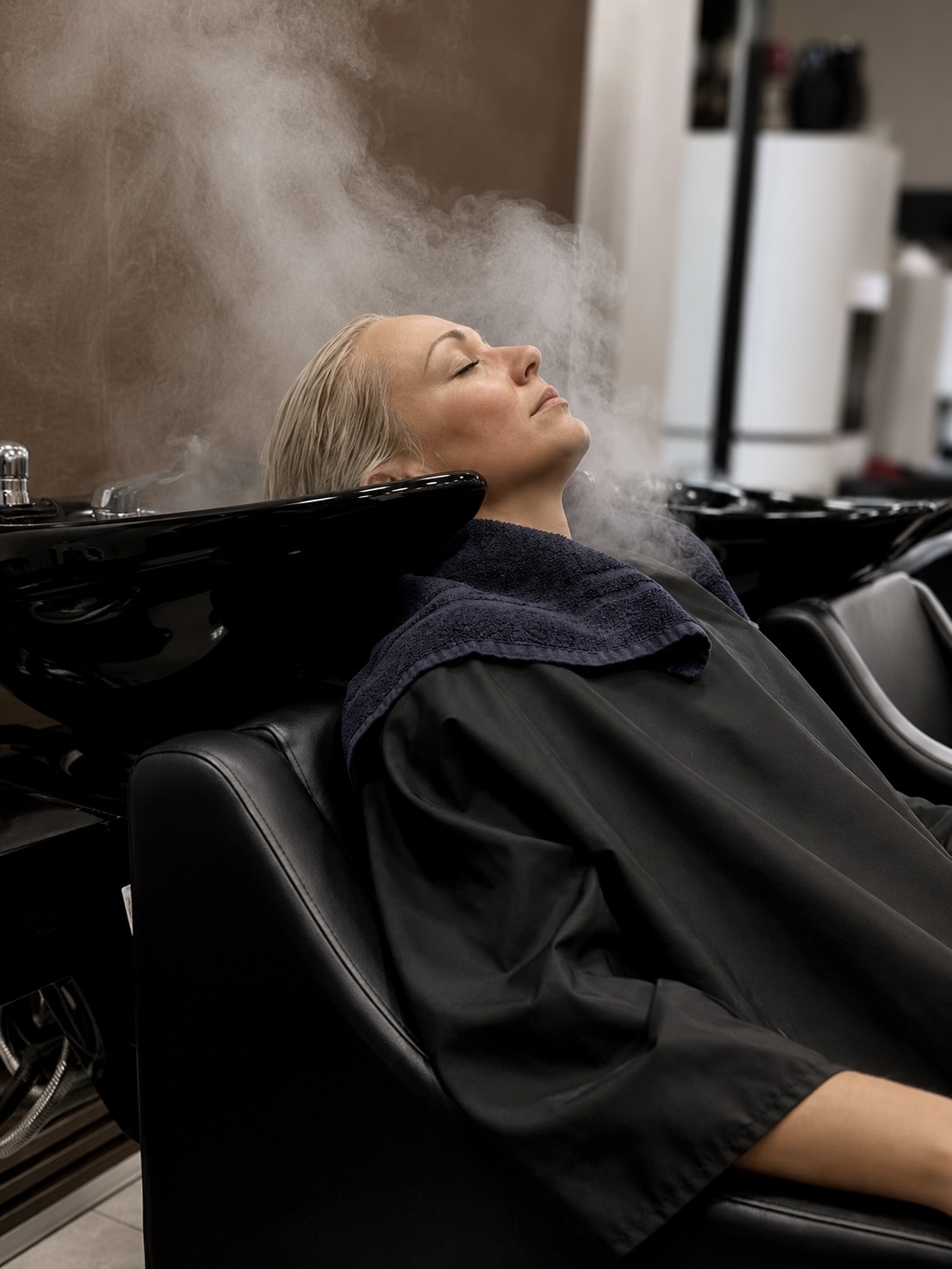 Woman with blonde hair reclines with head back in salon sink, steam rising from hair, wearing black cape and towel, salon background